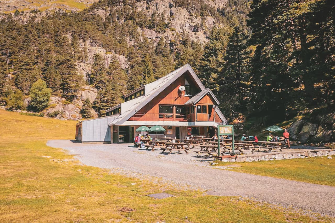 Le chalet du clot en bois, entouré de forêt et de falaises, avec une terrasse animée où des randonneurs se reposent au soleil.