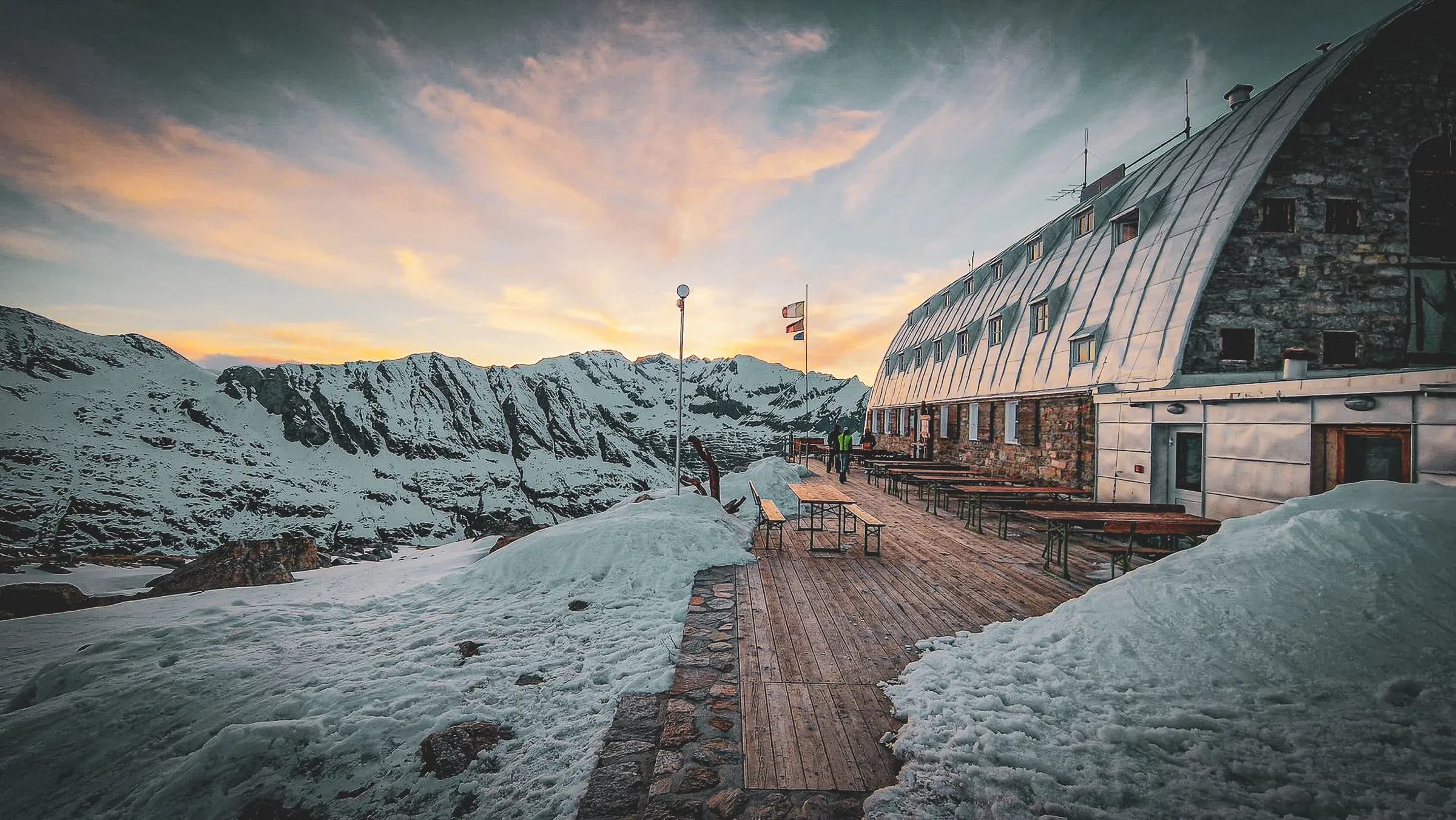A stone and metal mountain refuge at the top of an Alpine region, with a wooden platform in front, lined with snow. In the background, majestic mountains rise up, covered in snow, while