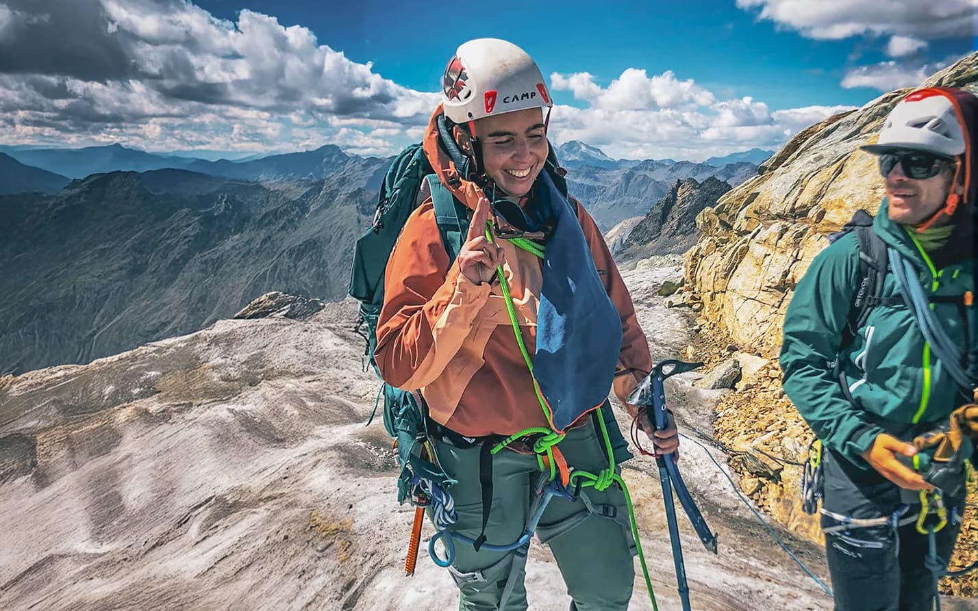 A mountaineer stands at the top of a mountain, smiling as he holds up a finger in victory. He is wearing a protective helmet and hiking gear, with a large rucksack and a coloured cord.