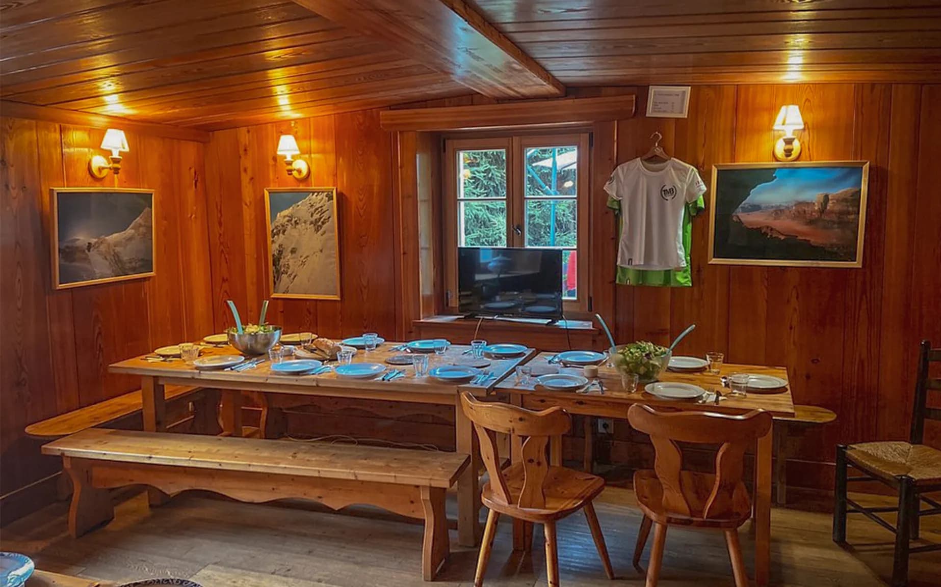 A warm wooden interior with wood-panelled walls, lit by wall lights. A large wooden table is set with blue plates, glasses and a salad bowl, ready for a meal.