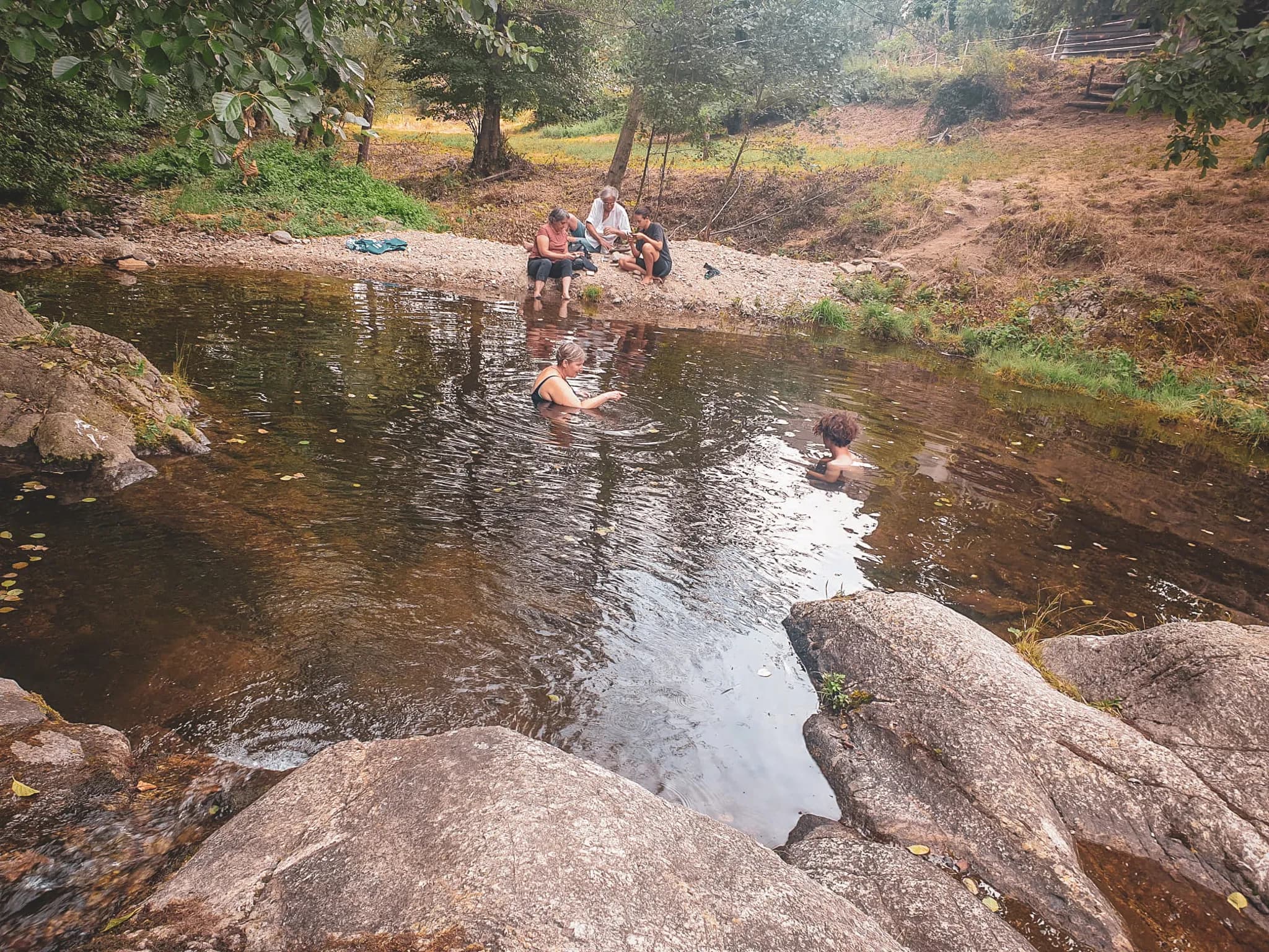 A group of people relaxing by a small, clear stretch of water, surrounded by nature. Two people are bathing in the water, while others are sitting on rocks or on the bank. The scene