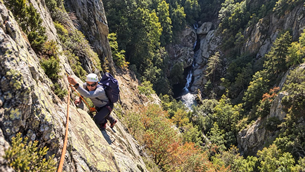 Een klimmer klimt tegen een rotswand op, boven een weelderig bos en een waterval.
