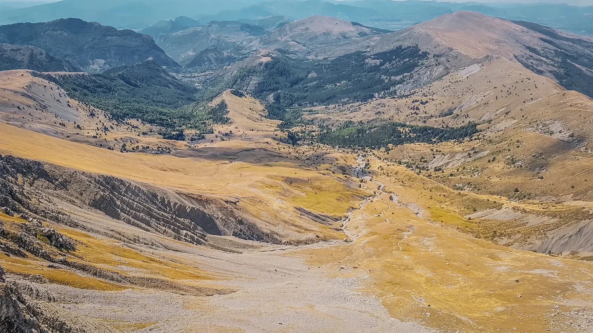 A vast, open mountain landscape, with hills and valleys. The slopes are covered with yellowed grass and rocky outcrops, while green areas can be seen in the distance. At the back