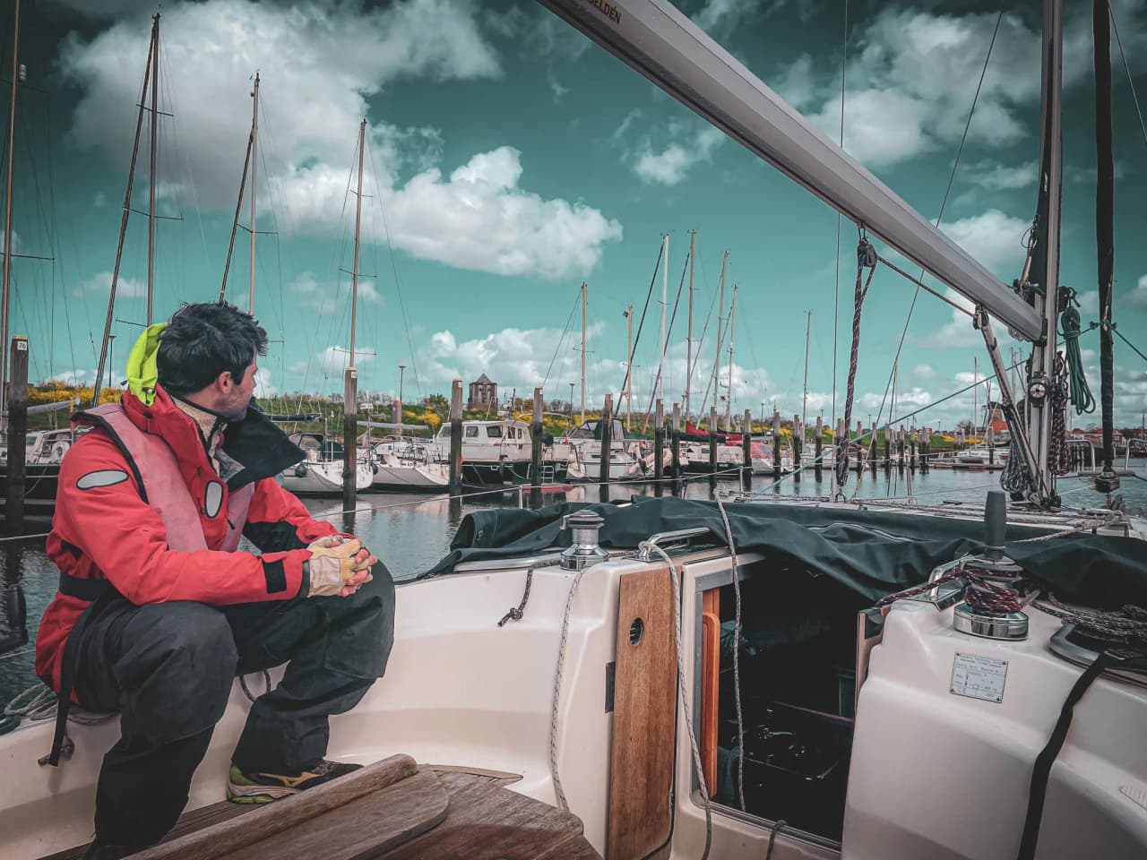 Een zeiler bekijkt de haven vanaf een zeilboot, onder een dramatische hemel met gestreken zeilen.