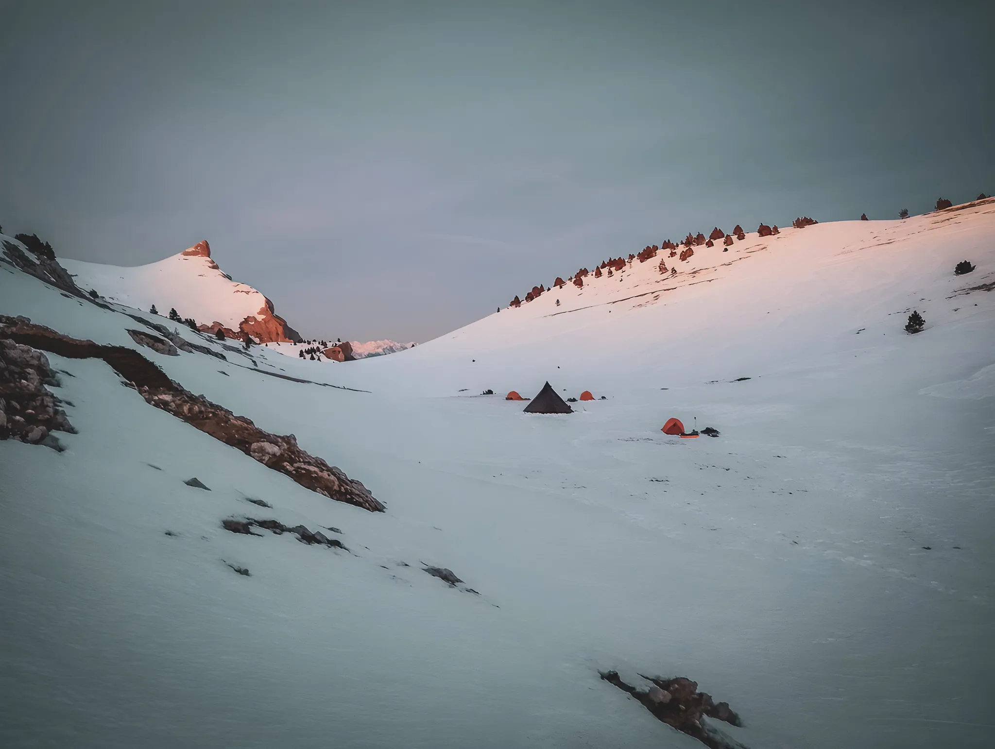 Bivak onder een pastelkleurige hemel, omringd door bergen met besneeuwde toppen en wilde landschappen.