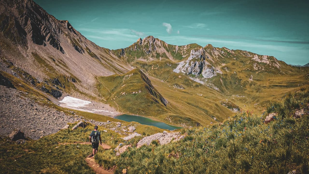Un randonneur sur un sentier montagneux verdoyant, avec un lac turquoise en arrière-plan.
