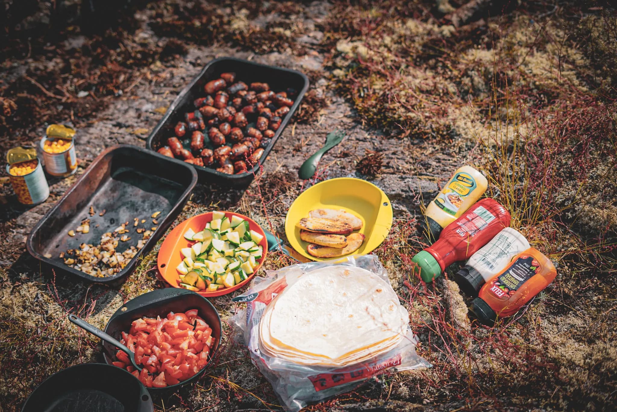 An outdoor food preparation area, with various ingredients spread out on the ground. There are grilled sausages in a black dish, diced courgettes in an orange dish, as well as