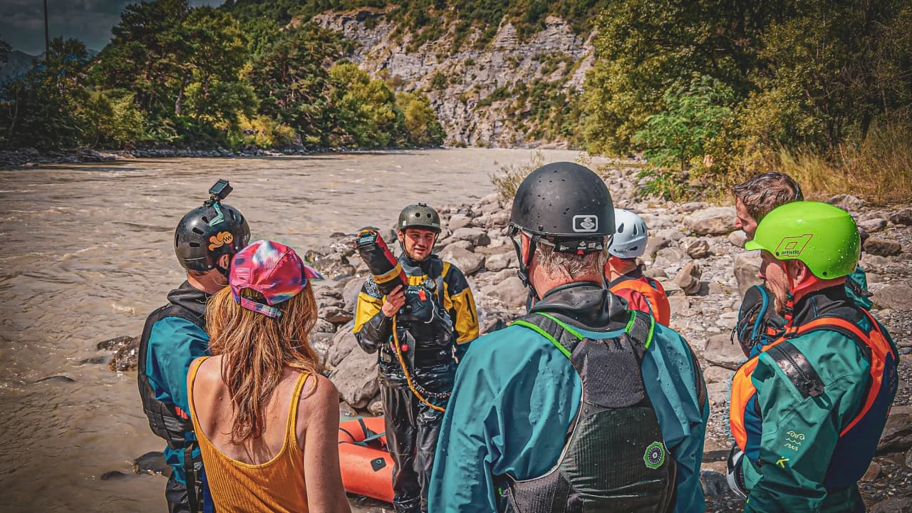 Een groep wandelaars die naar een instructeur luistert aan de oever van een groene alpenrivier.