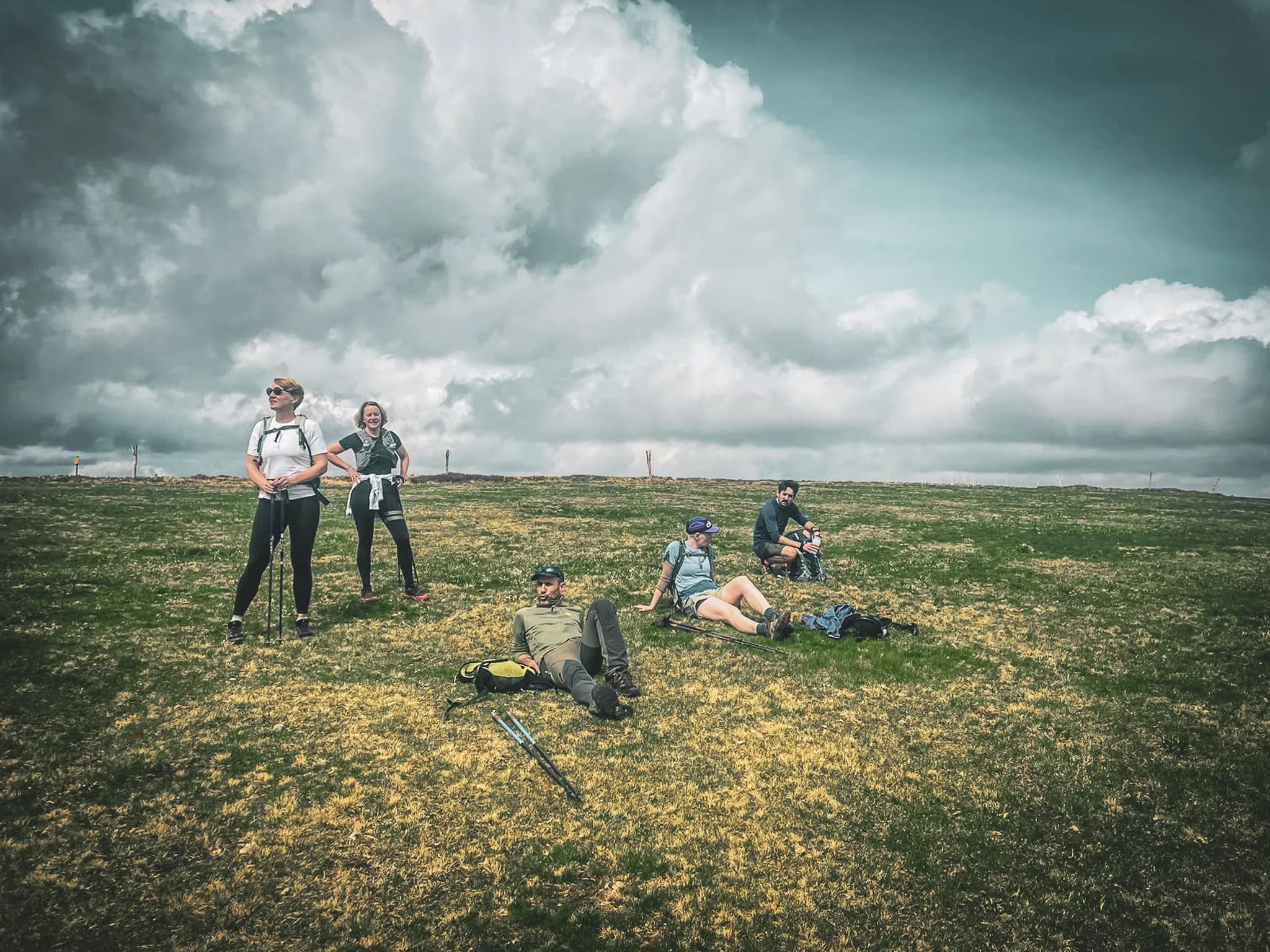 A group of walkers enjoying a break on a green hillside under cloudy skies.
