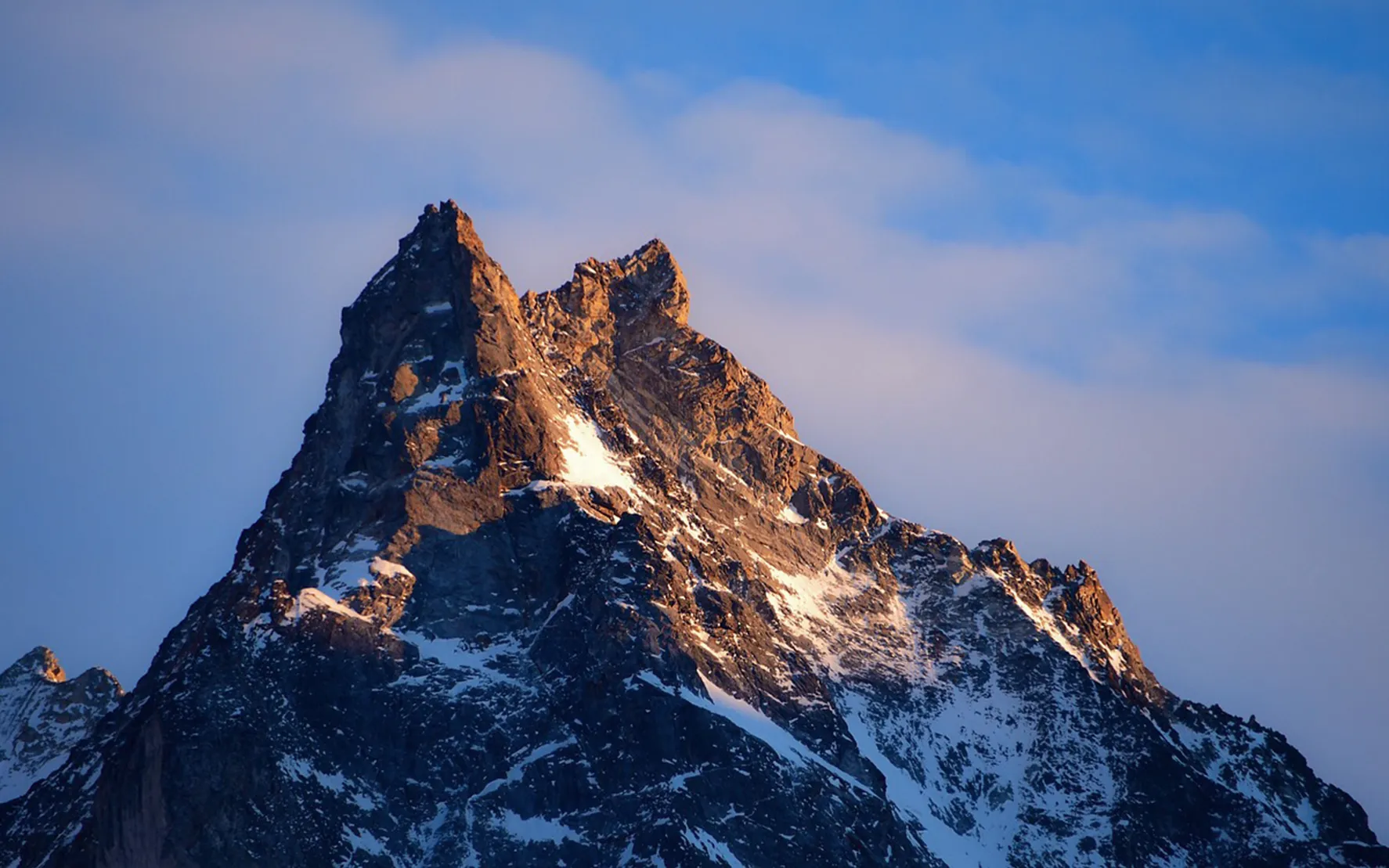 A majestic mountain peak, with craggy crags and eternal snow, stands proudly beneath a blue sky dotted with clouds.