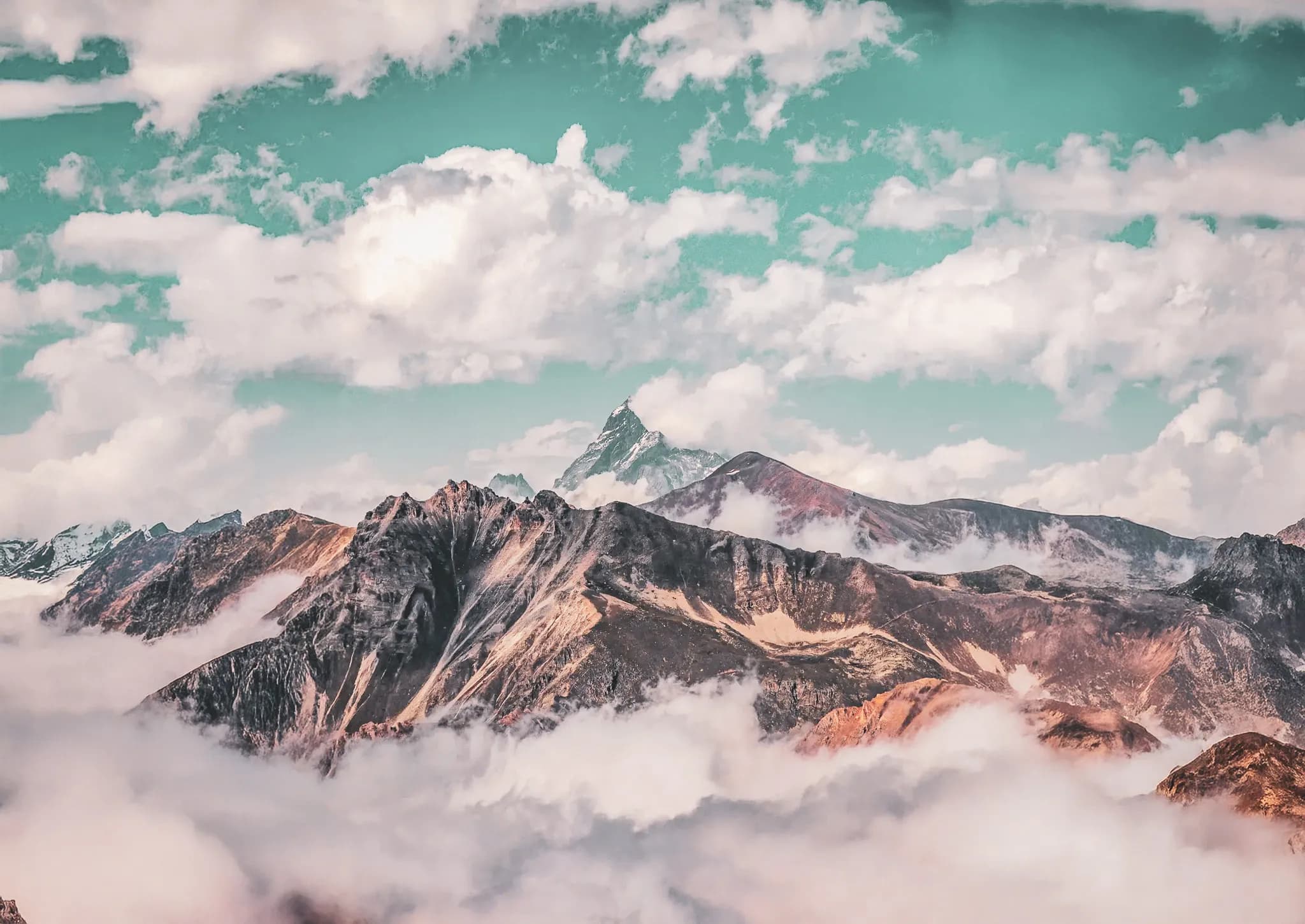 A majestic mountain landscape under a blue-green sky, dotted with white clouds. Craggy peaks rise above a sea of fog, while orange and brown rocky slopes are the backdrop for a spectacular view.