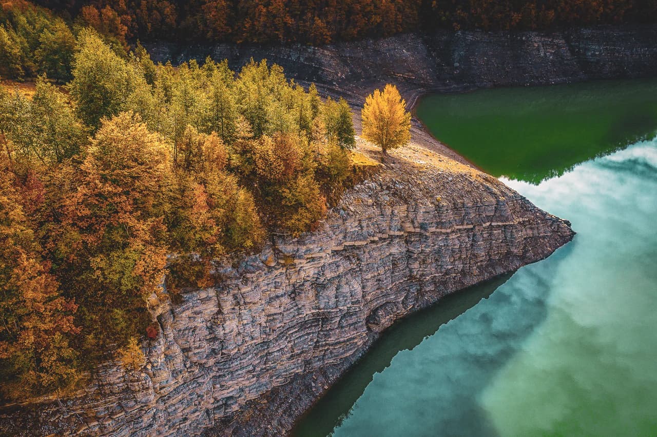 A natural landscape with a steep cliff overlooking a lake of green hues. The cliff is lined with trees in autumn colours ranging from orange to yellow.