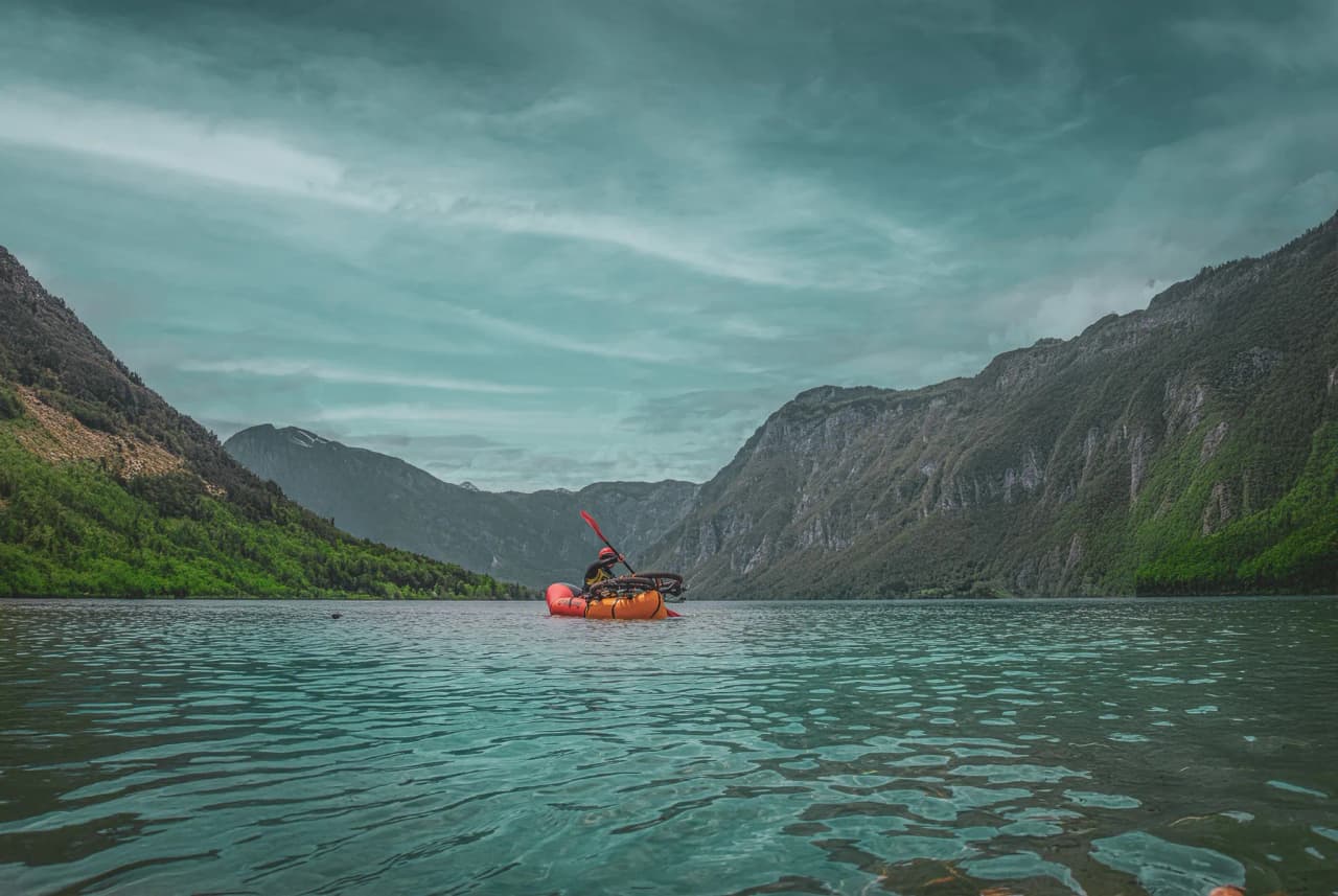 A calm lake surrounded by green mountains, with one person sitting in an orange kayak in the middle of the water. The sky is cloudy with shades of greyish blue, and the reflections on the surface of the water are very bright.
