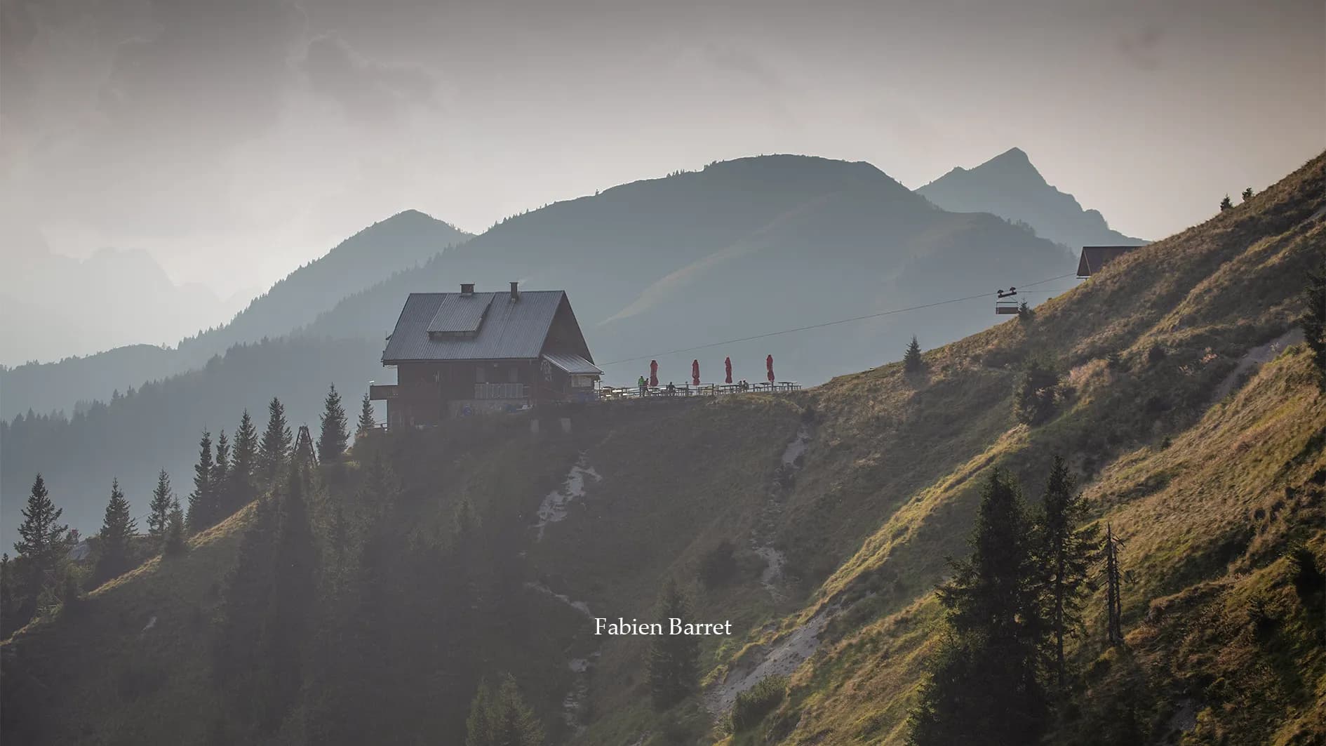 A wooden chalet stands on a verdant summit, surrounded by majestic mountains. The building, equipped with solar panels, is framed by a tranquil atmosphere at dusk. Winding paths surround