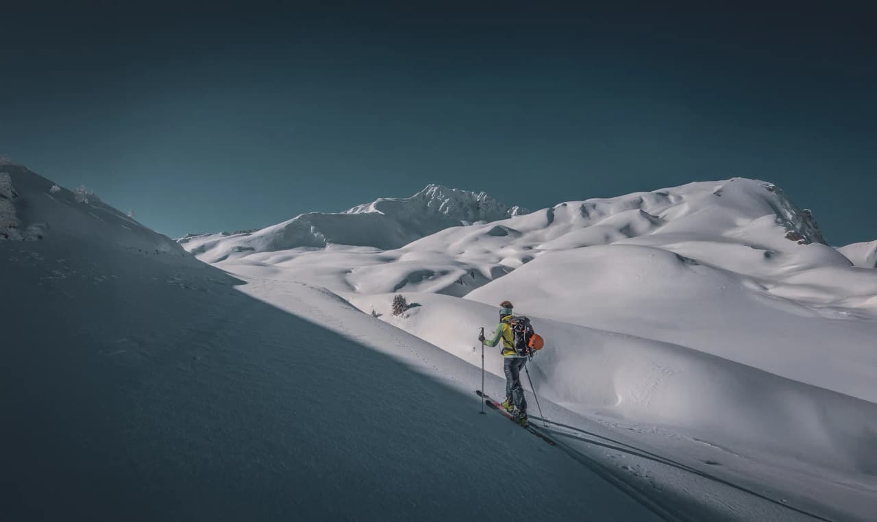 A skier ventures across a snow-covered mountain landscape, surrounded by rolling hills and majestic peaks. The sky is a deep blue, contrasting with the pure white of the snow.