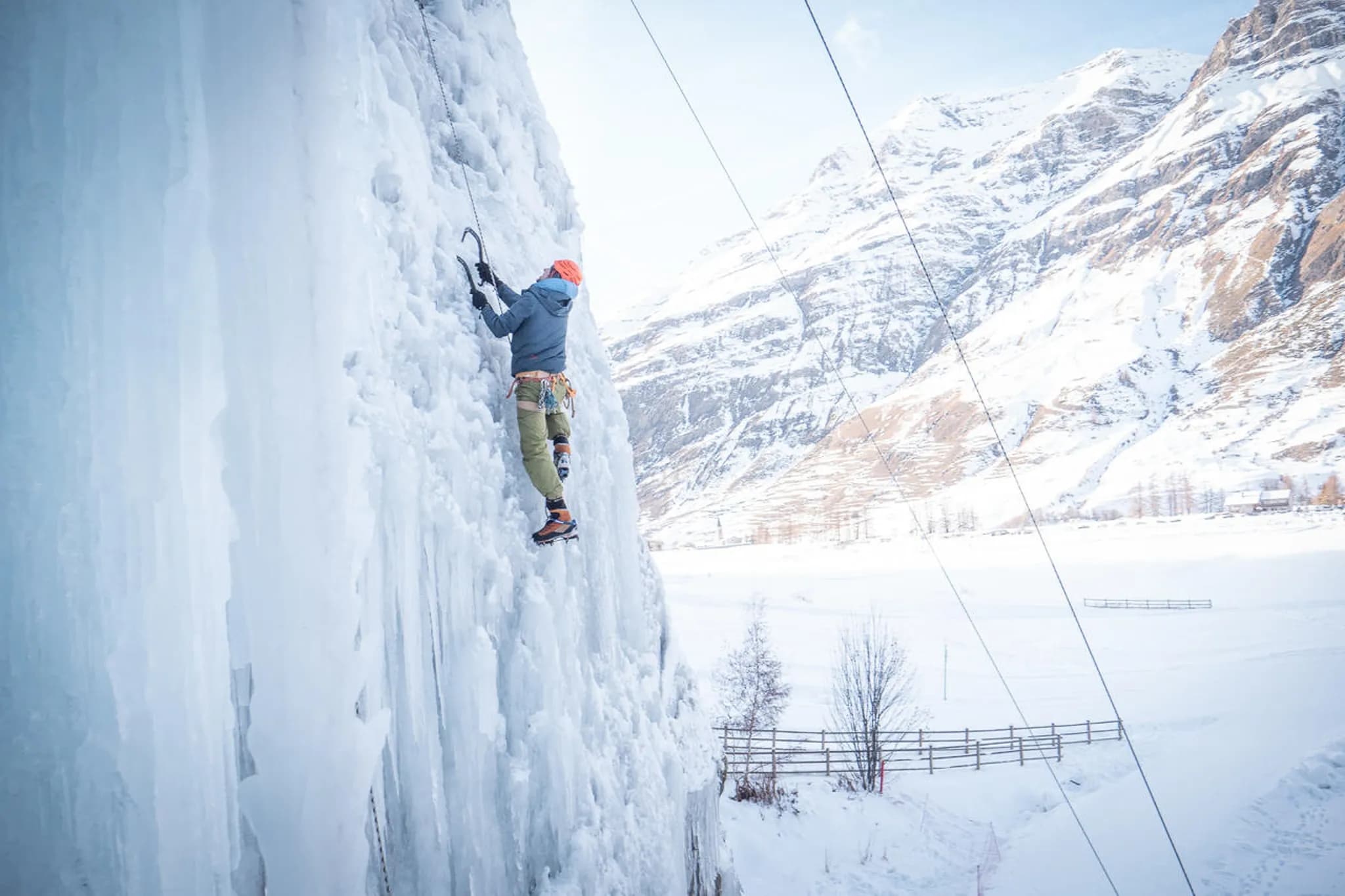 Un alpiniste grimpe une imposante cascade de glace, équipé de piolets et de crampons. Il porte une veste gris foncé et un bonnet orange, tandis que son harnais est attaché à une corde de sécurité. En