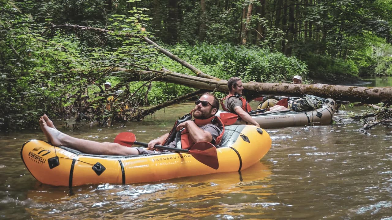 Paddlers relax in a packraft on a green river, surrounded by nature.
