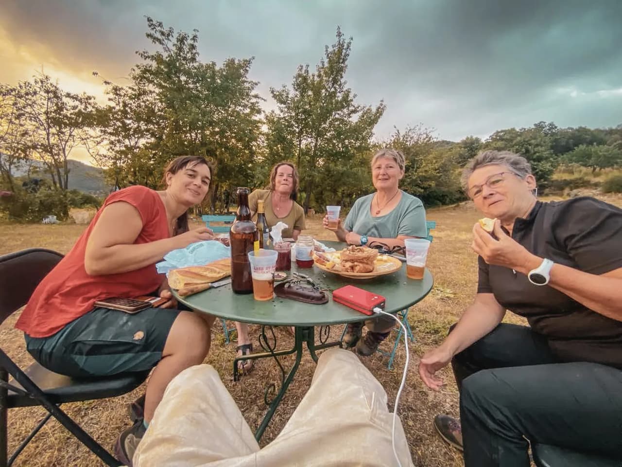 Vier lachende vrouwen rond een tafel, die een maaltijd delen op het platteland bij zonsondergang.