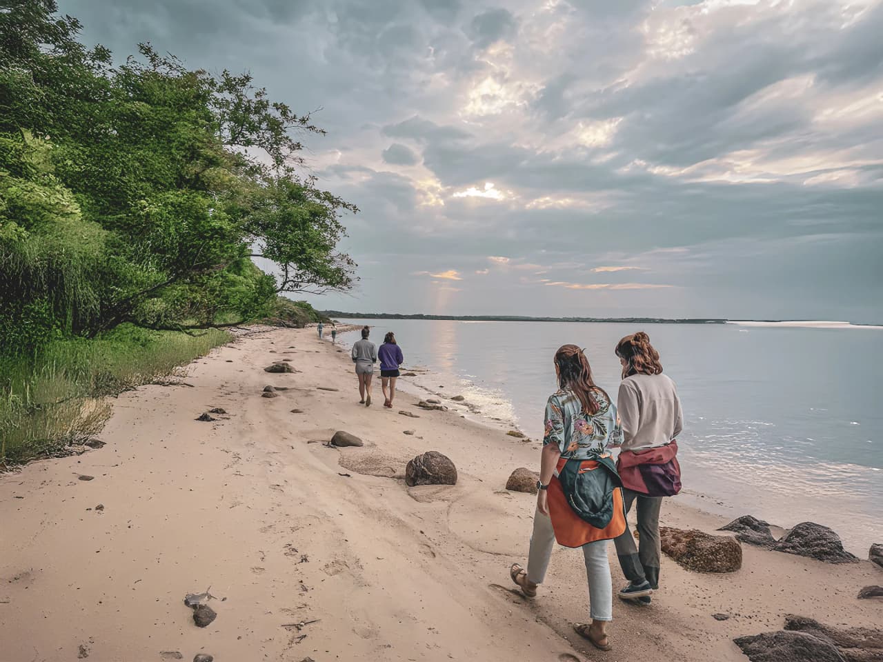 Deux personnes marchent sur une plage de sable, bordée de verdure, sous un ciel nuageux.