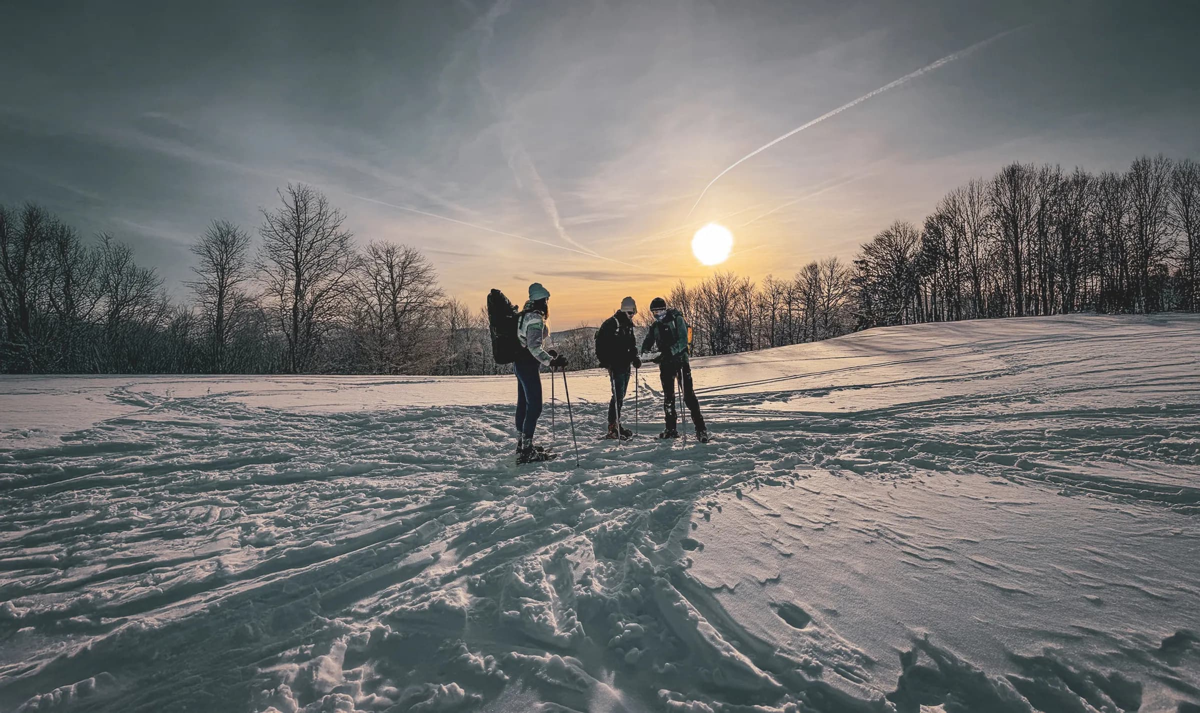 A group of three people, equipped with hiking poles, are standing on a vast field of snow. One of them, wearing a silver jacket and hat, seems to be chatting to the others, who are also dressed for the day.