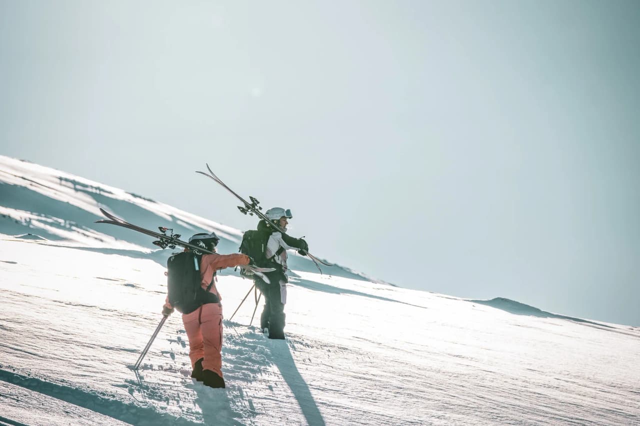 A pair of skiers stand on a snowy slope, with skis on their shoulders and rucksacks. One is wearing an orange jumpsuit, while the other is dressed in black and white. The ar