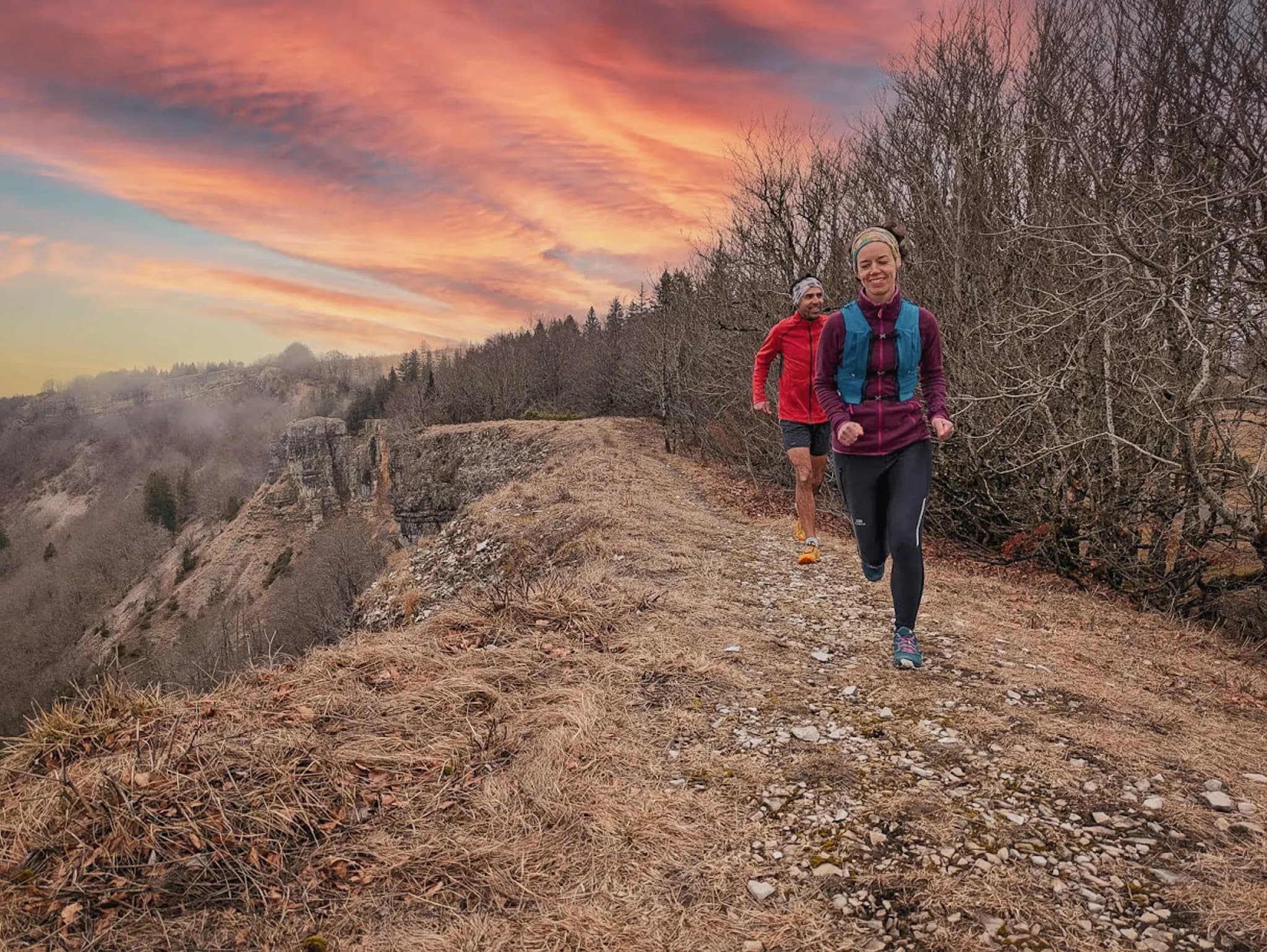 A nature trail with two runners in action. The first runner, a woman, smiles as she runs, wearing a light jacket and leggings, while the other, a man, follows her with a smile.