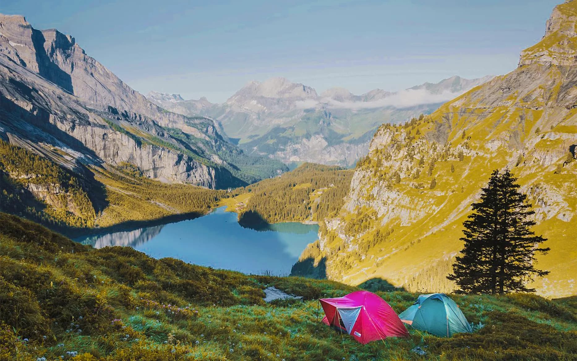 A spectacular mountain landscape with high peaks and a tranquil lake in the background. Two tents, one pink and one green, are set on a lush green lawn, surrounded by wild flowers. The slopes