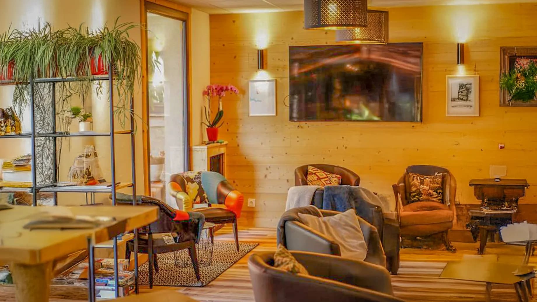 A welcoming living space with comfortable armchairs, wooden floors and a warm ambience. A large French window lets in natural light and offers a view of an outdoor patio. A piece of wooden furniture serves as a bookcase