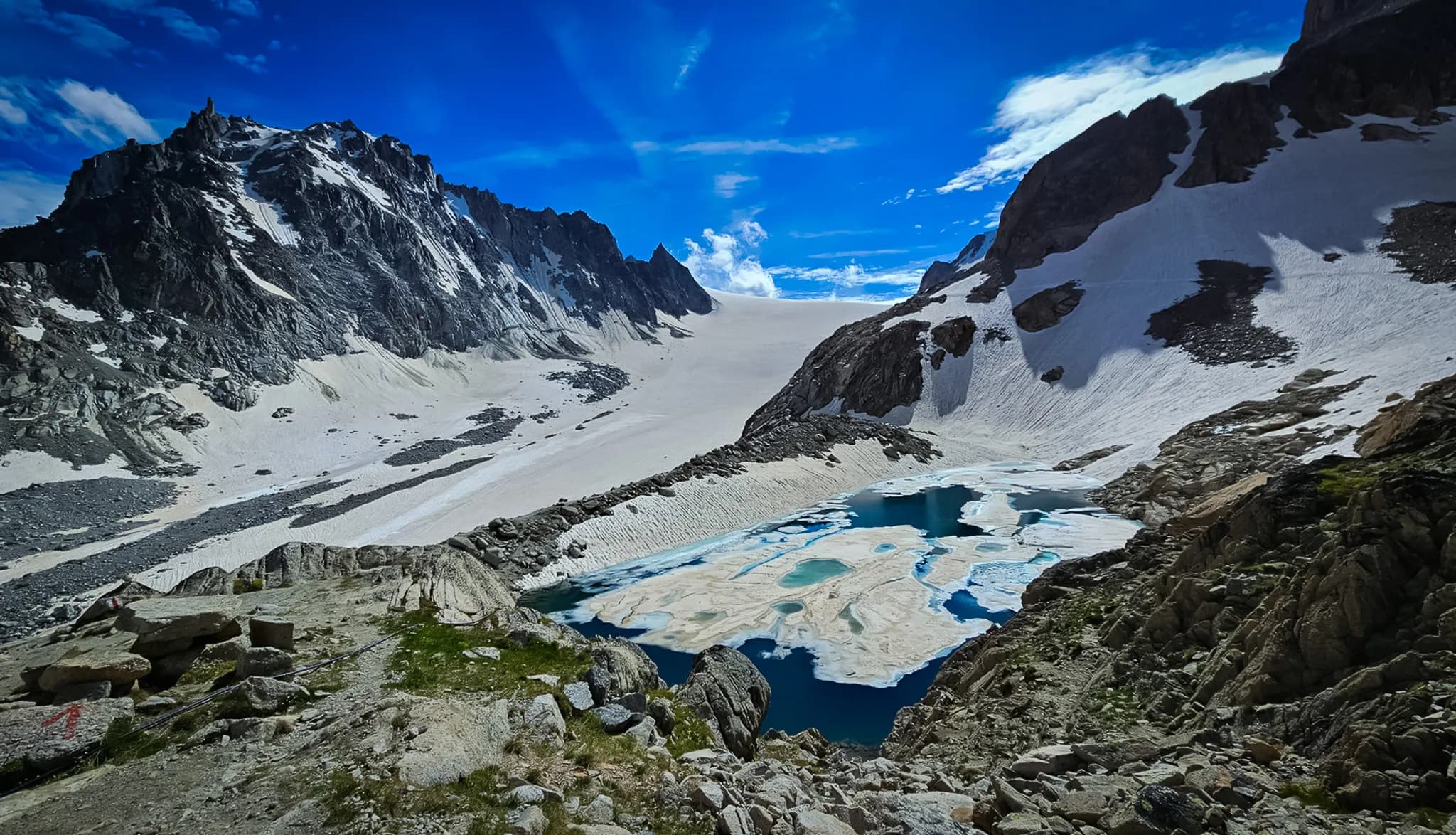 A spectacular mountain landscape with craggy, snow-covered peaks surrounding a turquoise lake. In the foreground, rocky and grassy terrain contrasts with floating areas of ice on the