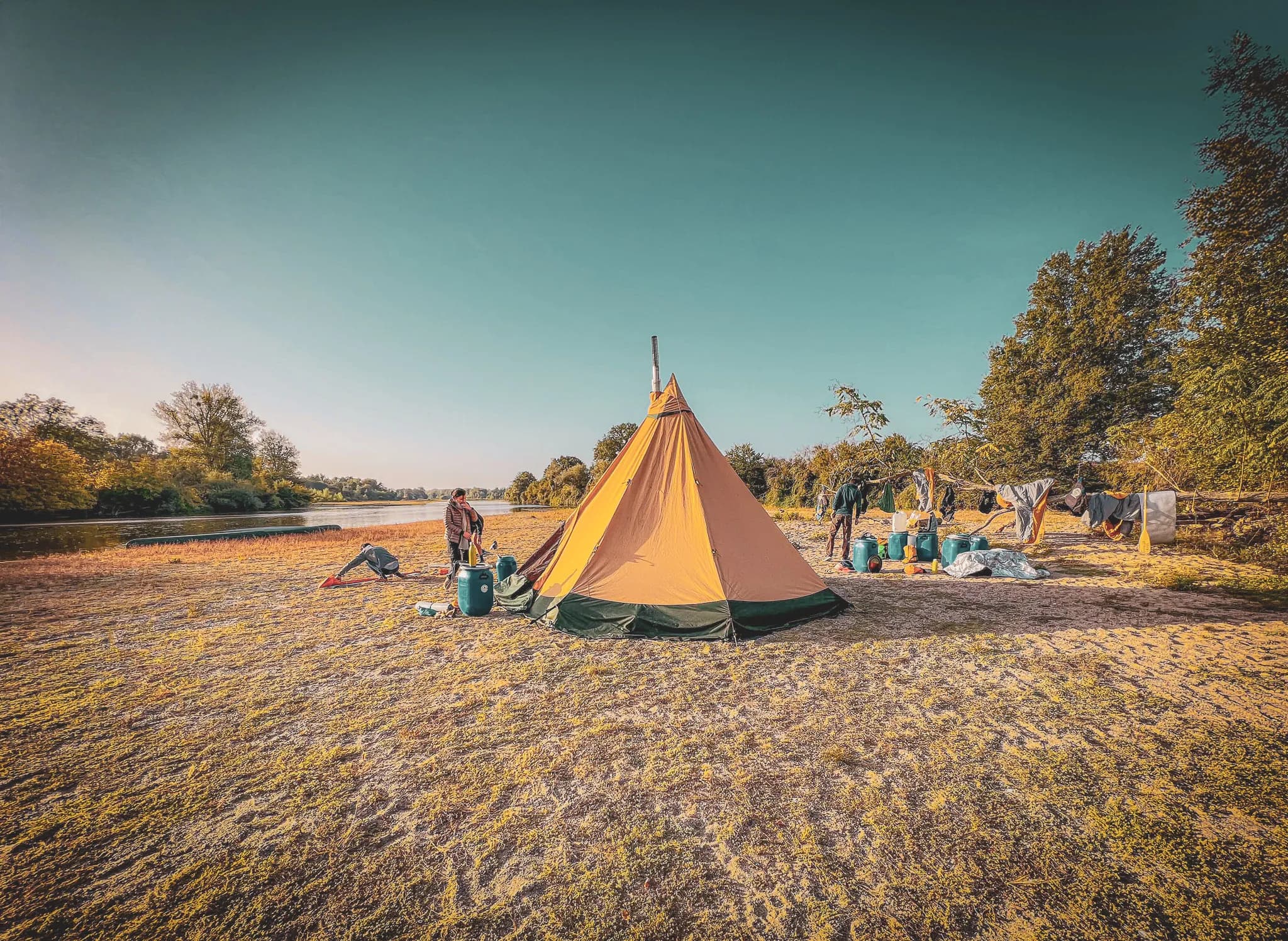 Un campement situé au bord d'une rivière, comprenant une grande tente en forme de cône de couleur orange. Des personnes sont visibles, l'une manipulant un équipement près de la tente, tandis que d'autres s'affairent autour. Des