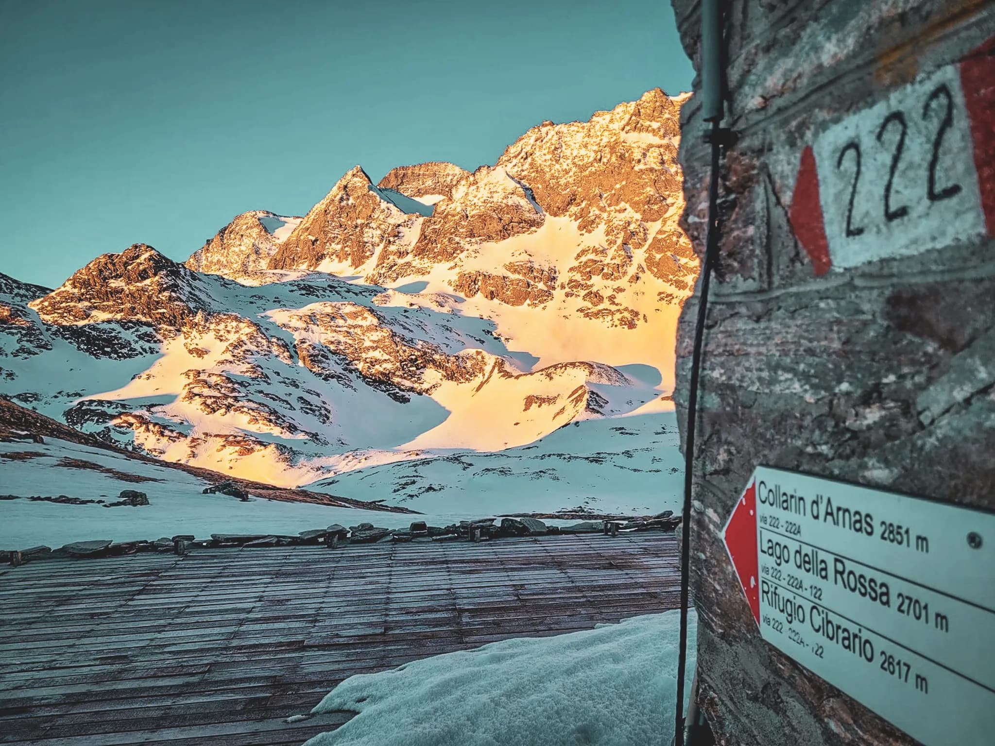 A spectacular Alpine landscape with majestic snow-capped mountains lit up by the morning sun. In the foreground, a well-kept wooden floor and directional signs indicating altitudes