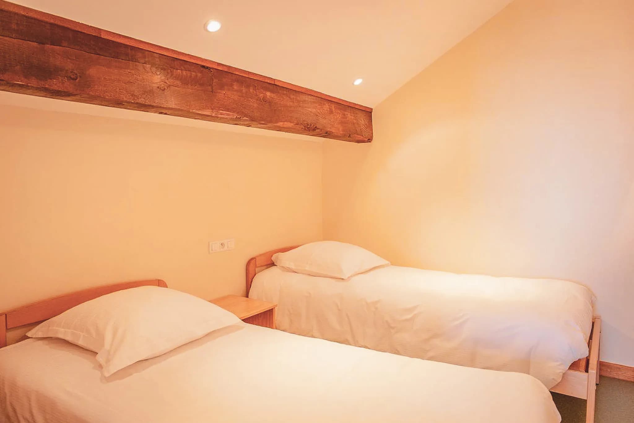 A well-lit room with two separate single beds. The beds are covered with white sheets and large pillows. A beige wall forms the backdrop, and a decorative wooden beam can be seen on the ceiling.