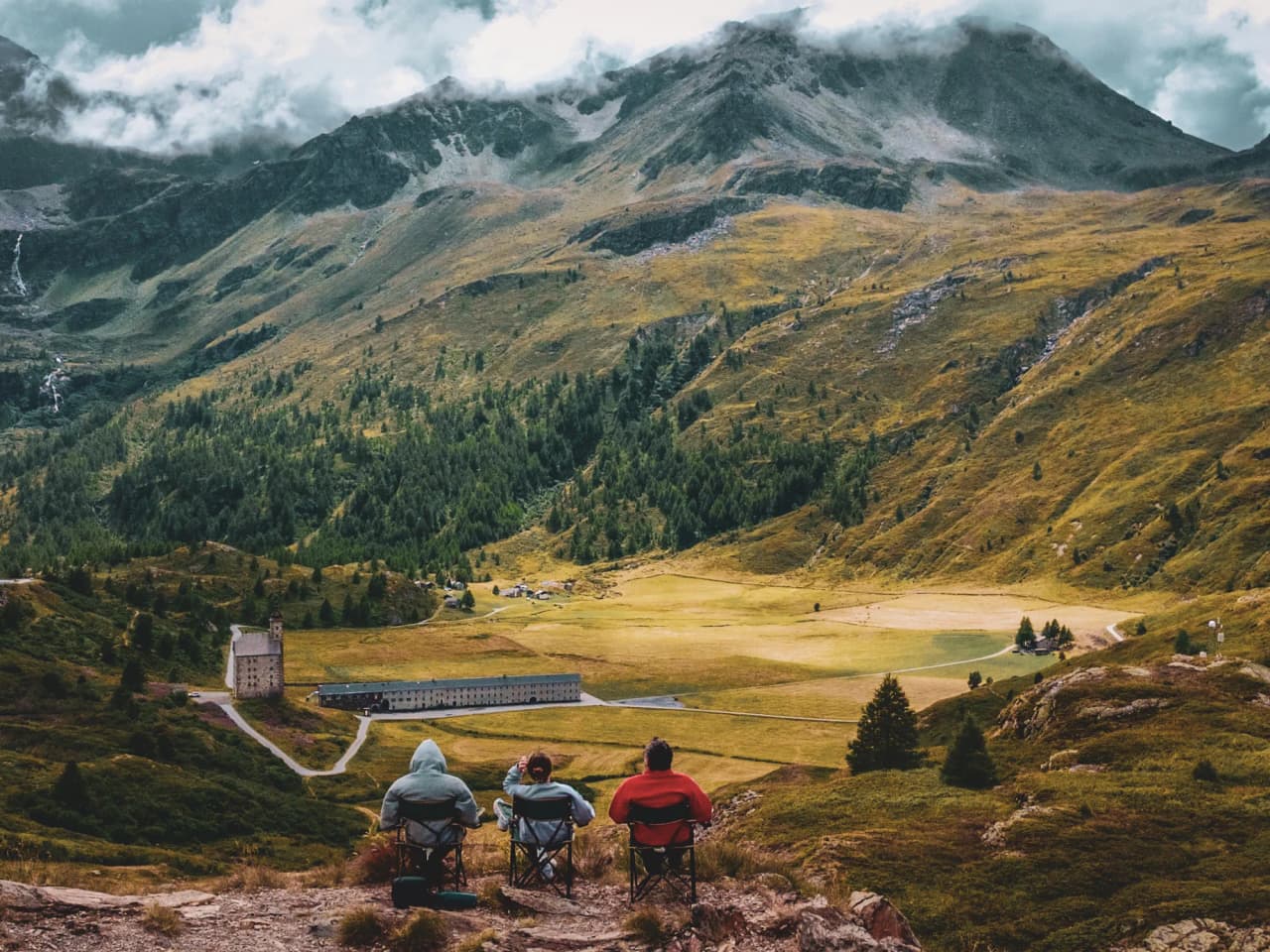 Three hikers admiring a vast mountain landscape in the Dolomites, resting on folding chairs.
