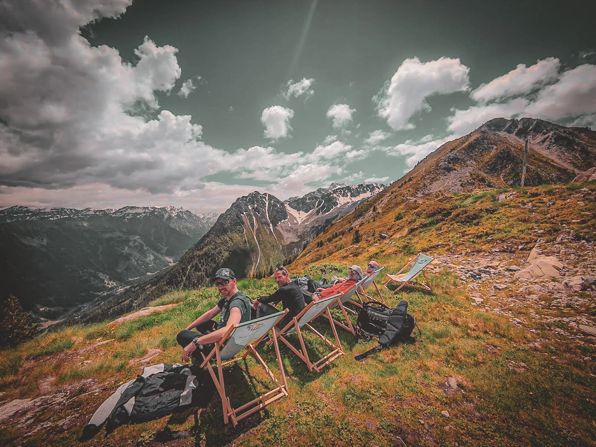 Four friends relax on chairs in a spectacular Alpine landscape, under a cloudy sky.