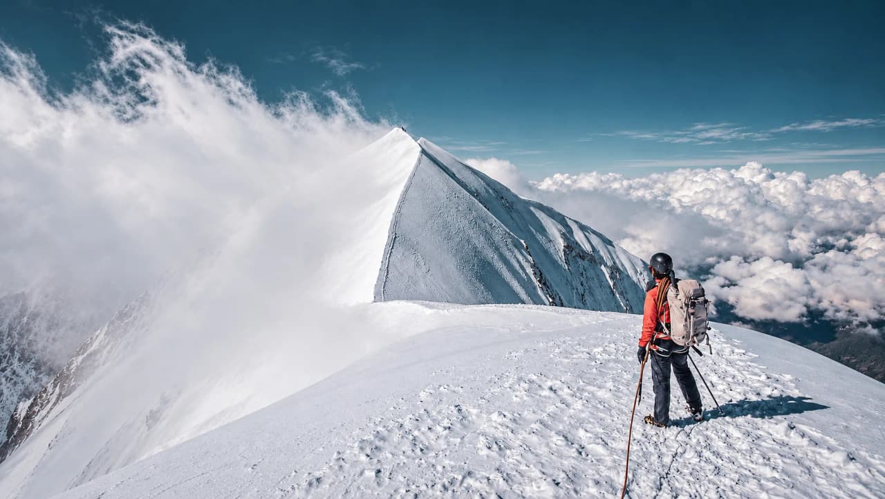 Een wandelaar op een besneeuwde bergtop, met uitzicht op majestueuze bergen onder een blauwe hemel.