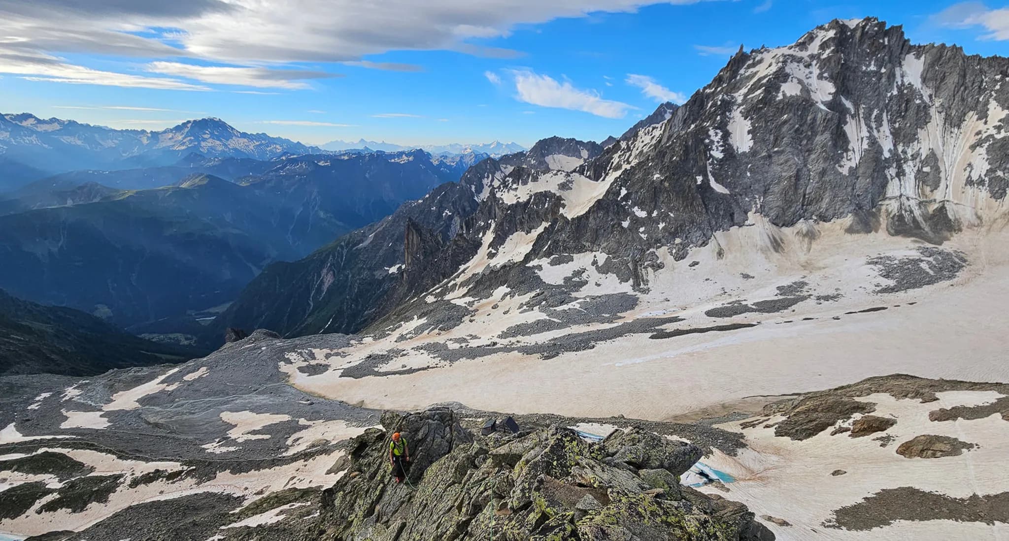 A spectacular mountain landscape with steep, snow-capped peaks, offering panoramic views of the valley below. The scene shows an equipped mountaineer making his way over rocky terrain, while
