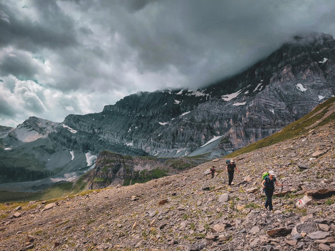 Three hikers climb rocky terrain, surrounded by majestic Alpine mountains, under a cloudy sky.