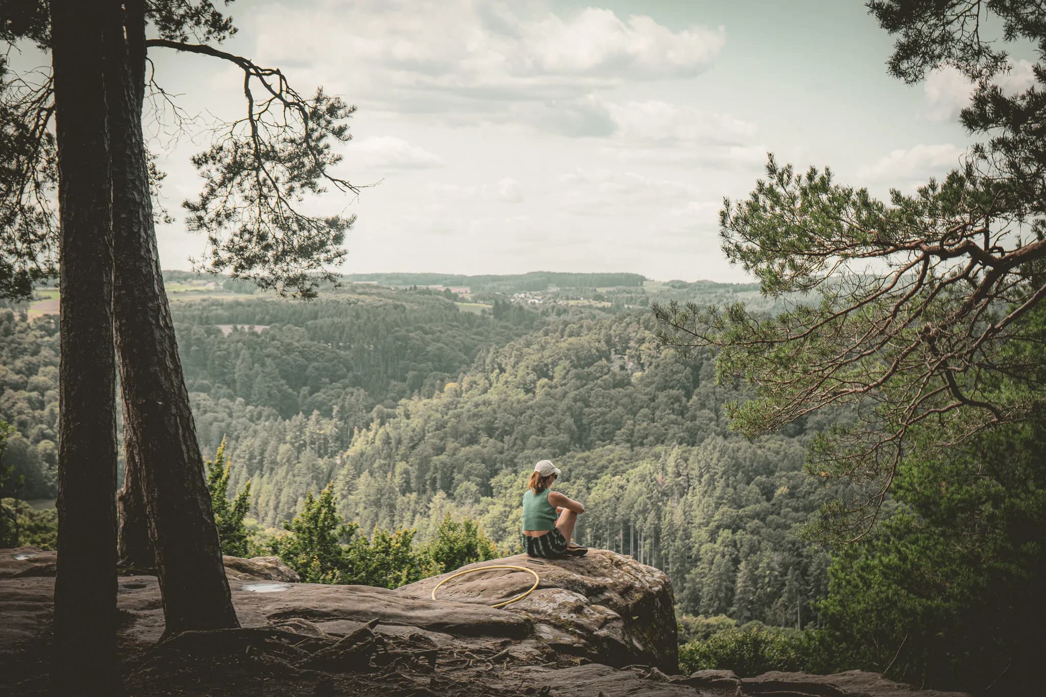 a young person sitting on a rock, gazing out at green forests under a cloudy sky.