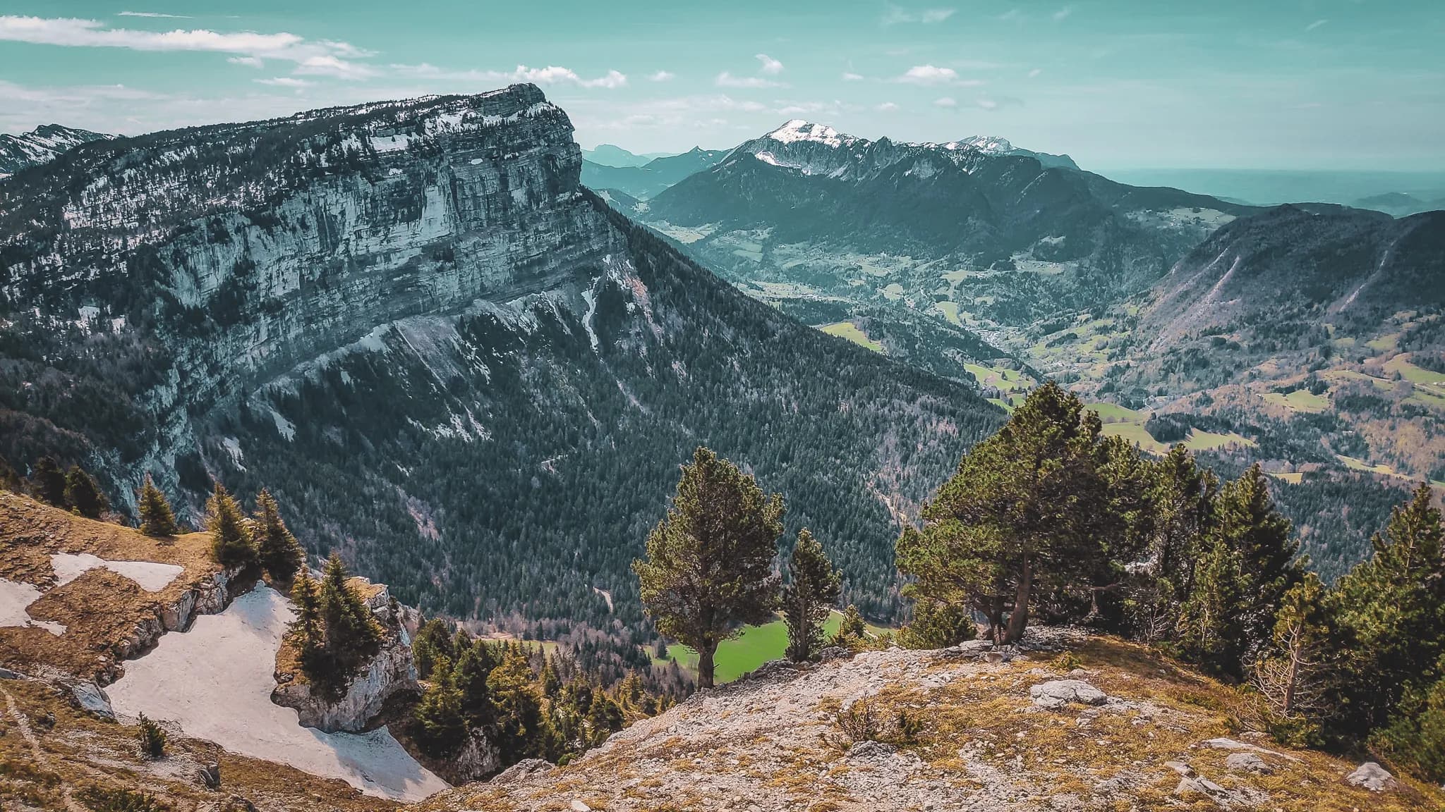 Un paysage montagnard spectaculaire, avec des sommets enneigés en arrière-plan. À gauche, une grande falaise escarpée domine la scène, tandis qu'à droite, des vallées verdoyantes s'étendent, parsemées