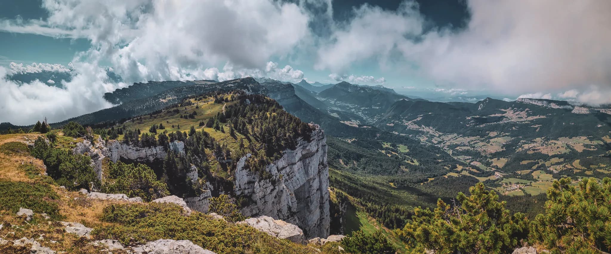 Un panorama époustouflant sur des montagnes verdoyantes, avec des falaises abruptes et des sommets majestueux entourés de nuages. La scène est caractérisée par une vaste étendue de forêts denses, des dunes de