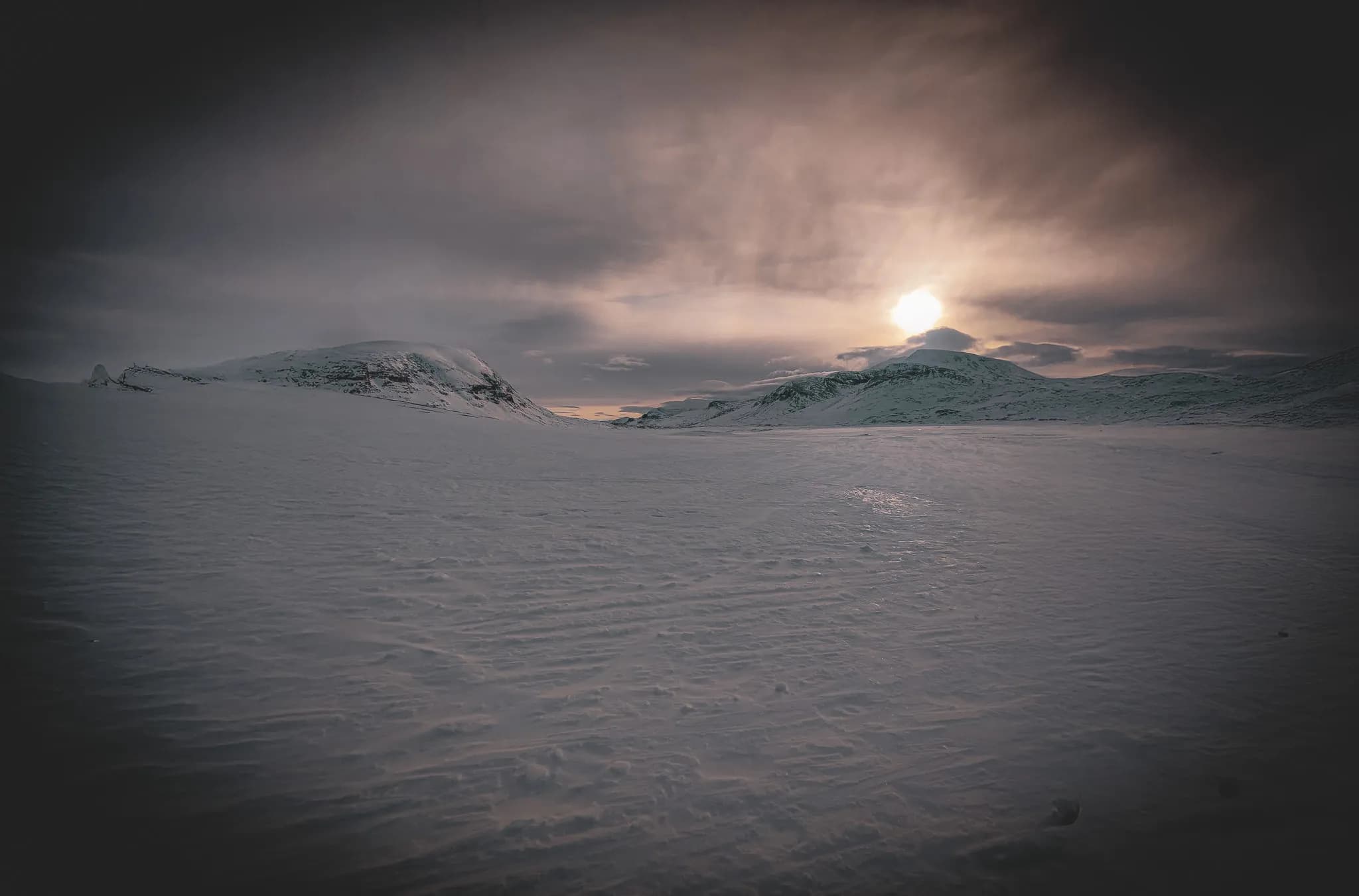 A snow-covered landscape unfolds under a cloudy sky, with sunlight piercing through the clouds to create a mysterious atmosphere. The snow covers the ground, highlighting the wave patterns.