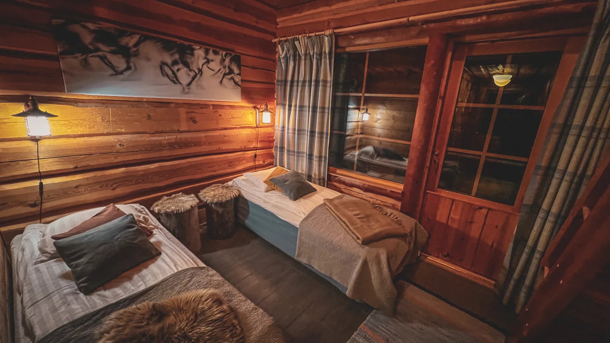 A cosy wooden bedroom, with log walls and warm lighting. Two single beds are furnished with colourful cushions and soft blankets. On the left, a hanging lamp lights up the space, while on the right, the light from the ceiling lights up the room.