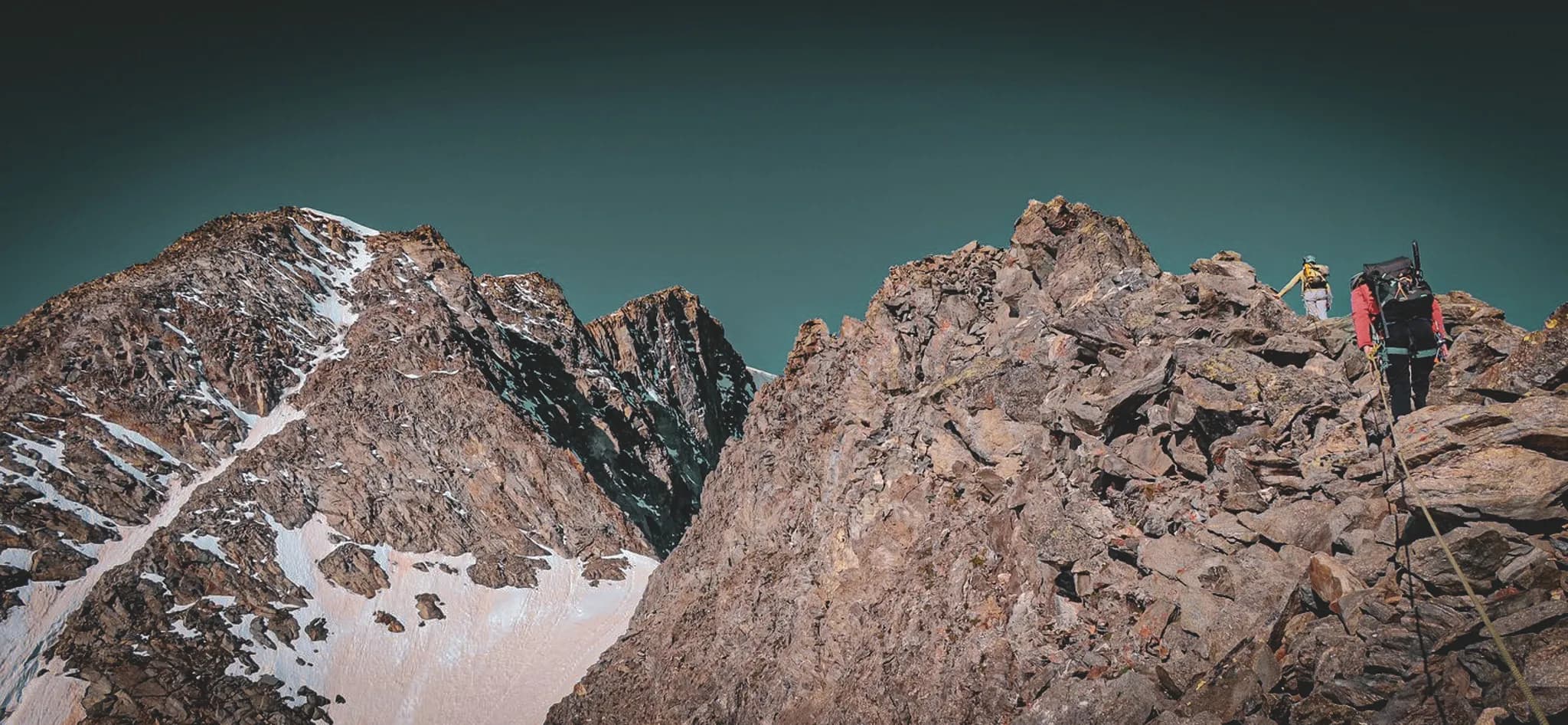 An impressive mountain landscape with craggy peaks covered in snow and rock. In the background, majestic ridges rise up against a deep blue sky. In the foreground