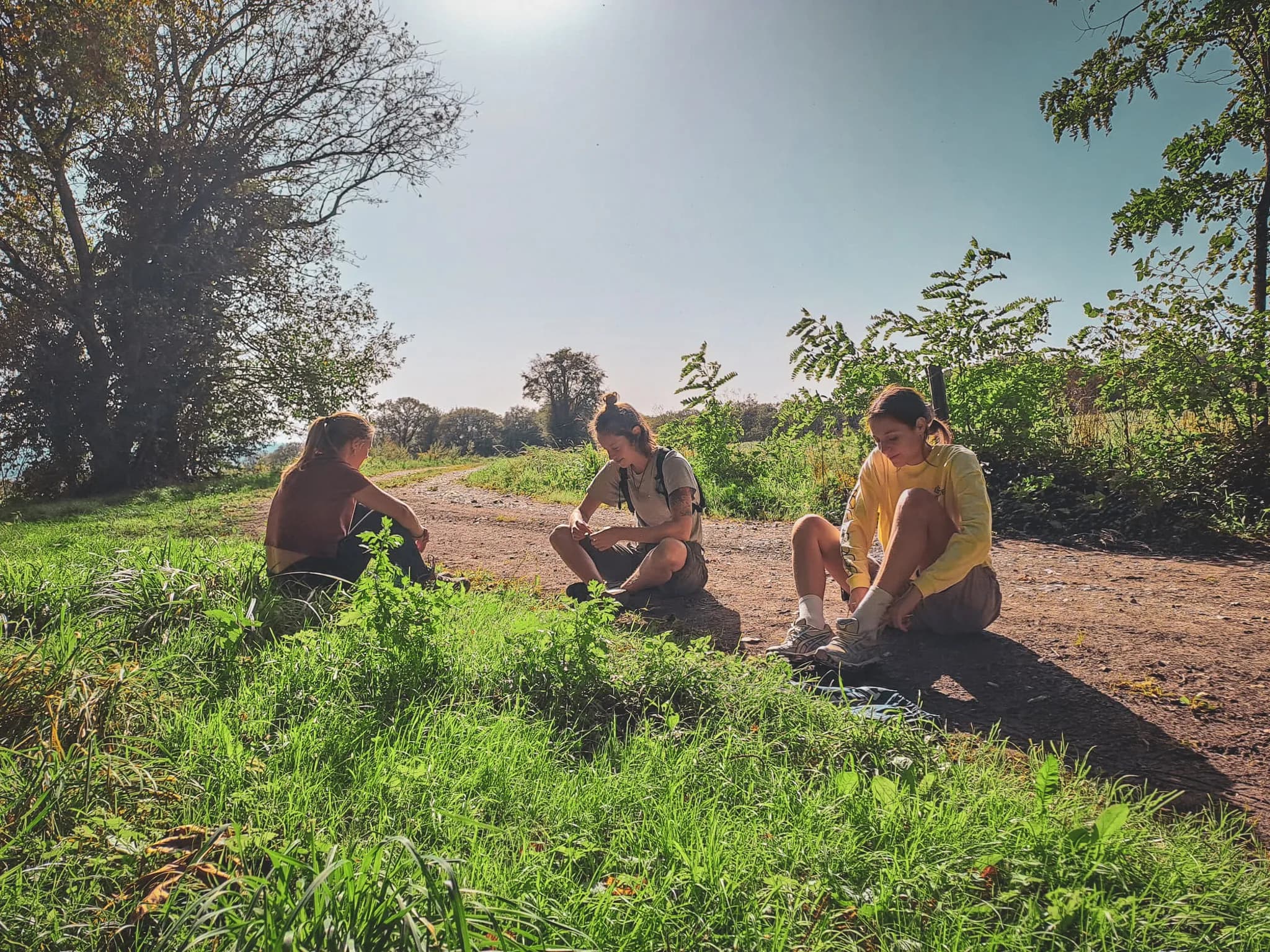 Three people sitting on a dirt path, surrounded by greenery. Two of them are leaning towards the ground, having a relaxed moment, while the third, wearing a yellow t-shirt, is waiting for the sun to rise.