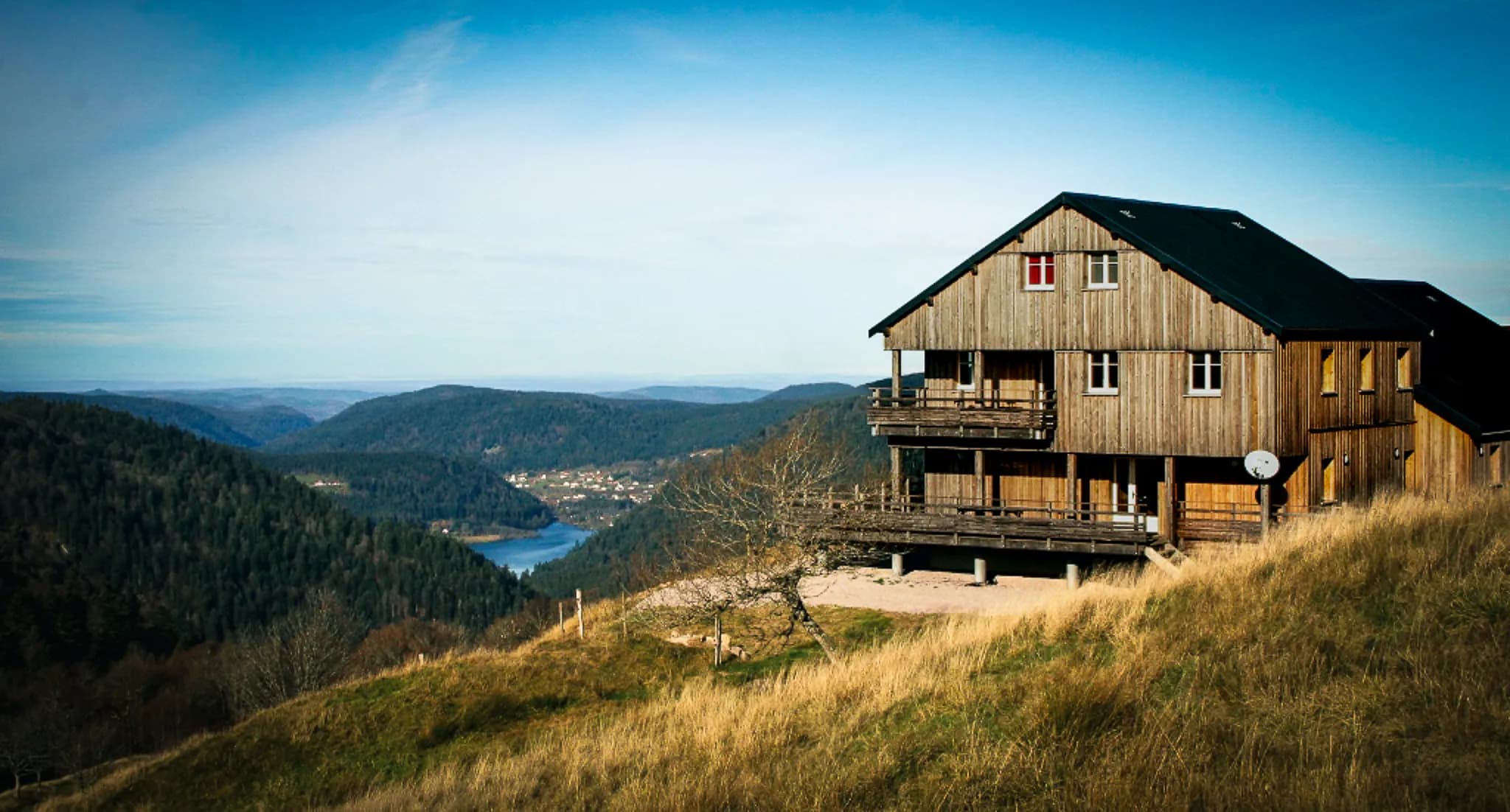A wooden chalet is set on a hill overlooking a green valley and a lake. The chalet has several storeys, with a wooden balcony running the length of the chalet. In the distance, mountains and forests