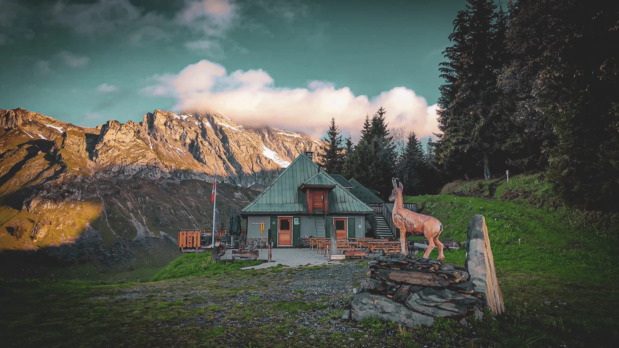 Un chalet en montagne avec un toit en pente, entouré de verdure et de grands arbres. Des montagnes majestueuses avec des neiges persistantes se dressent en arrière-plan, tandis que des nuages flottent dans un ciel bleu.