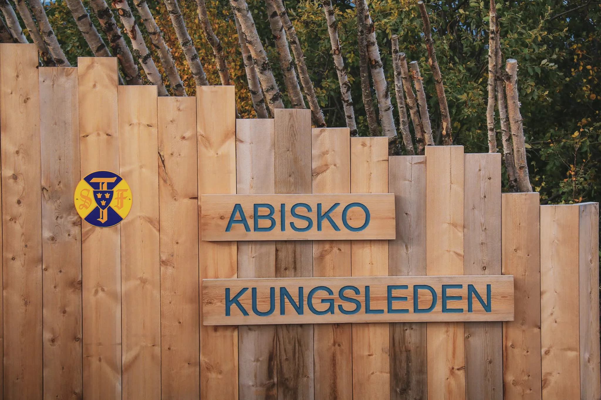 A stylised wooden panel features the inscriptions 'ABISKO' and 'KUNGSLEDEN' in blue letters, surrounded by wooden planks in a variety of colours. To the left, a circular arb