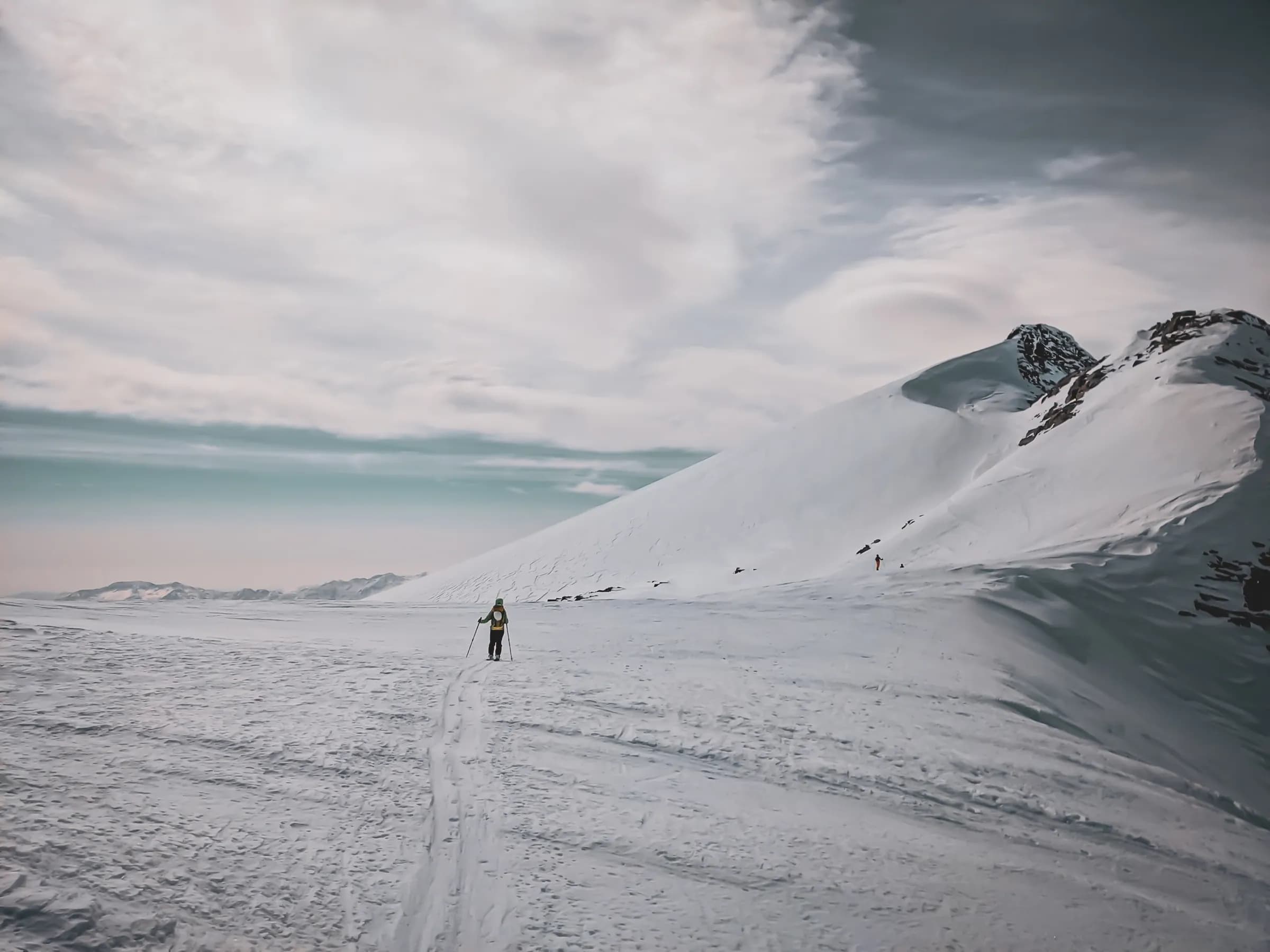 A snow-covered landscape, with a lone skier making his way across a surface of immaculate snow. In the background, a snow-covered mountain rises majestically, while the slightly cloudy sky