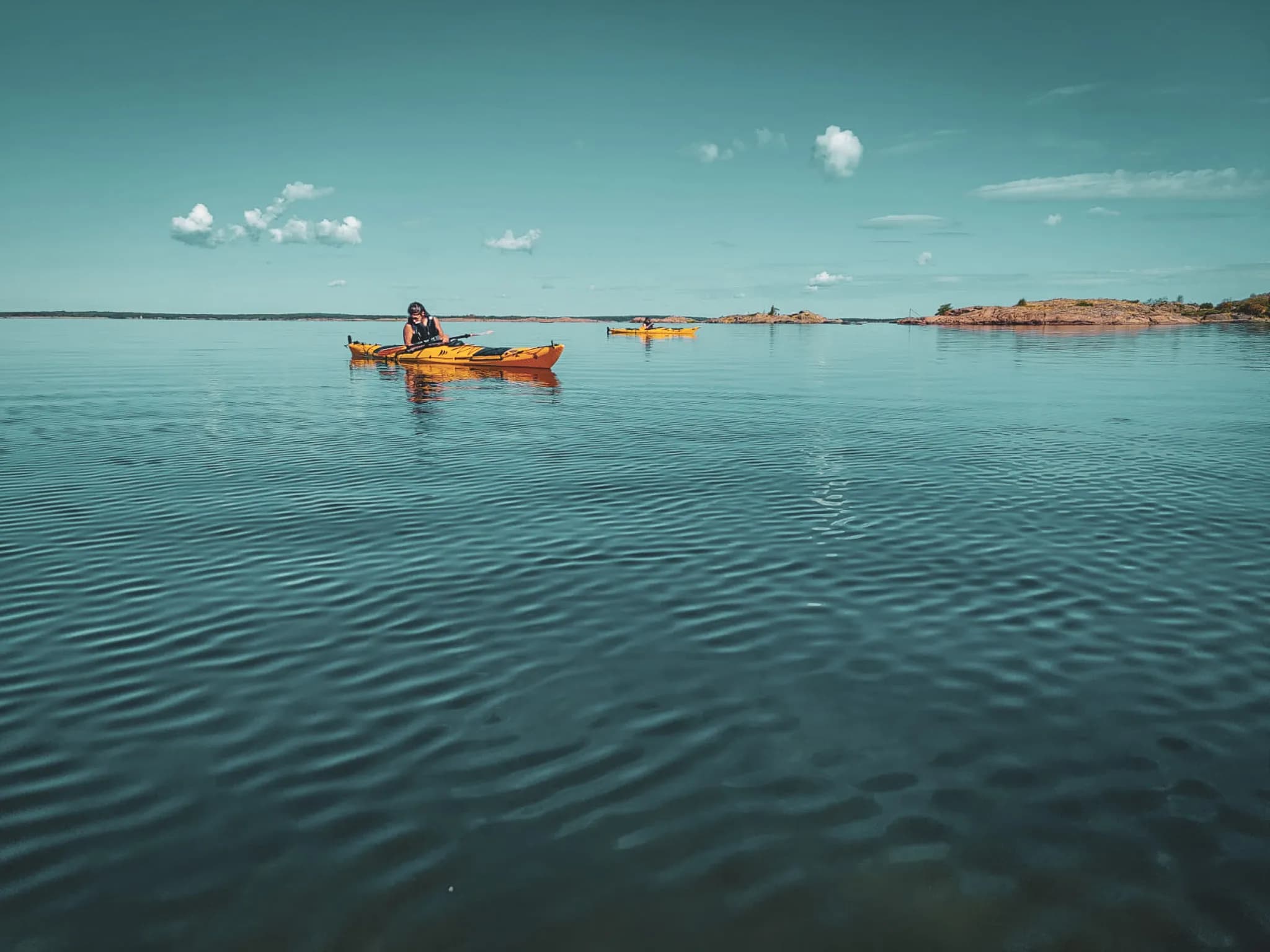 A kayaker glides quietly over a smooth surface of water, while other kayaks are in the background. The sky is clear with a few clouds, and in the distance, rocky islands emerge from the sea.