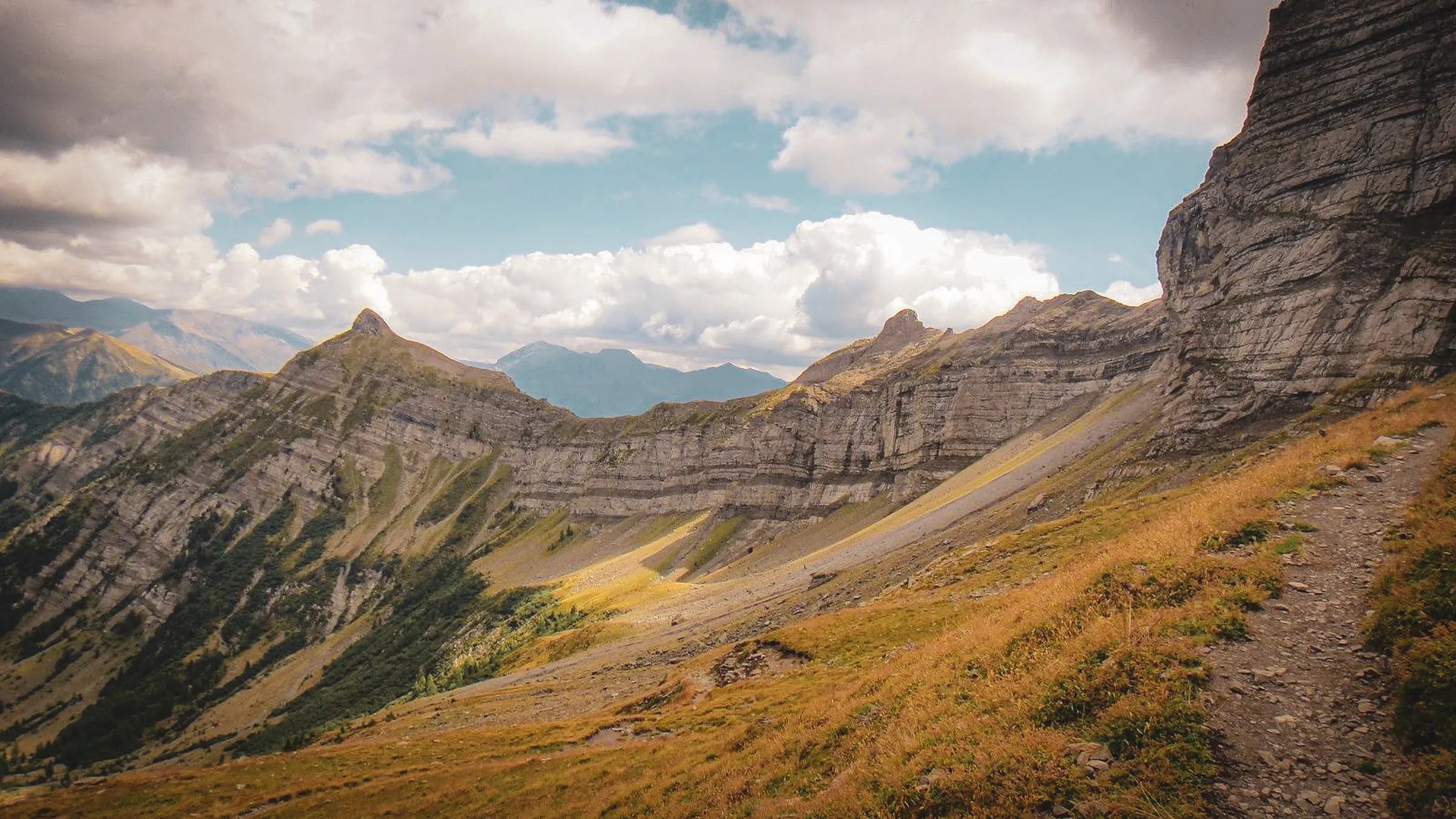 A spectacular mountain landscape with steep peaks and well-defined geological strata. Vegetation is particularly prominent in sunny areas, while grass and earth form a winding path.